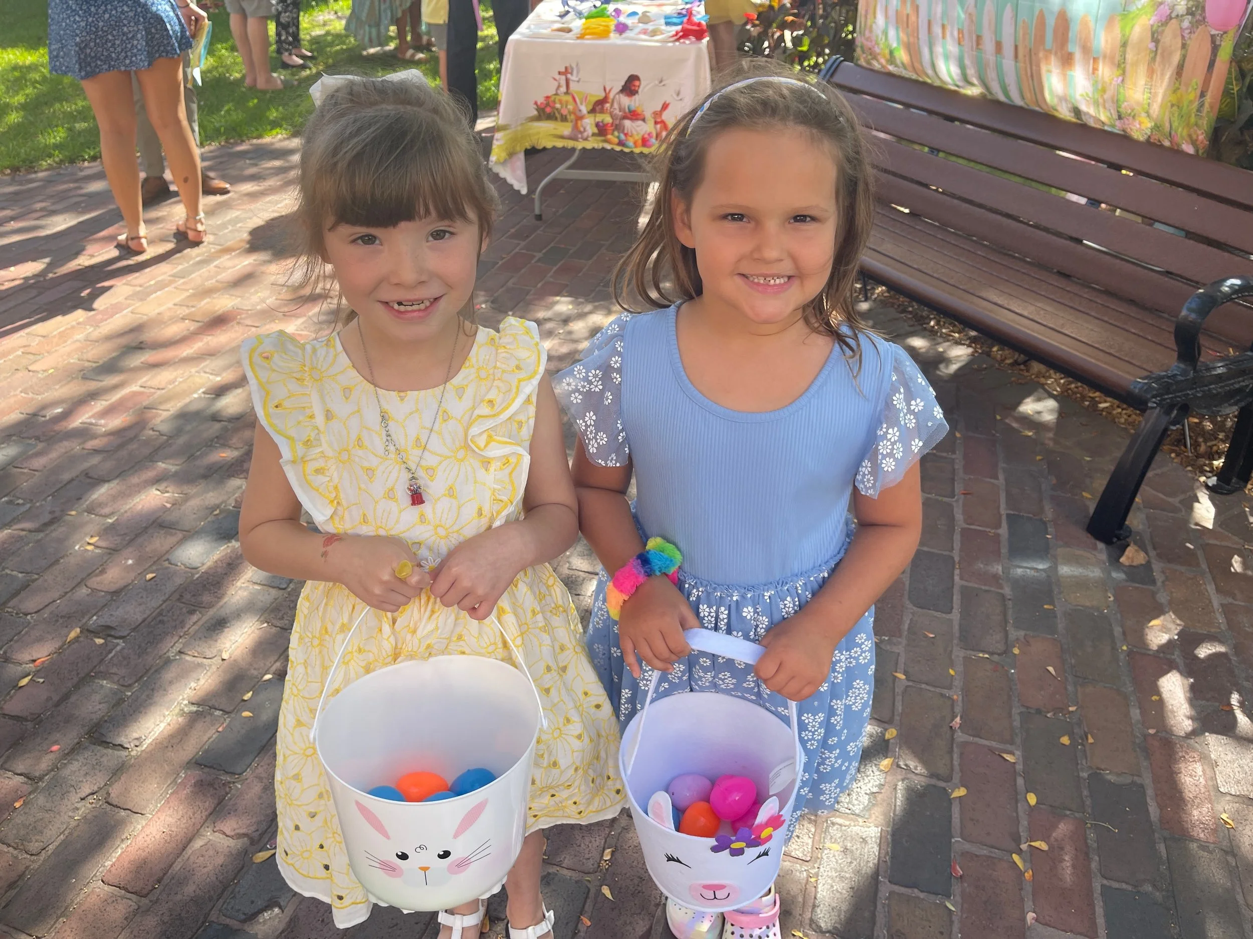 Two young girls standing outdoors on a brick pathway, smiling and holding white Easter basket buckets filled with colorful plastic eggs. One girl is wearing a yellow dress with floral embroidery and the other is in a blue dress with white floral details. In the background, people are gathered, and a table with a cloth featuring Easter-themed decorations is visible.