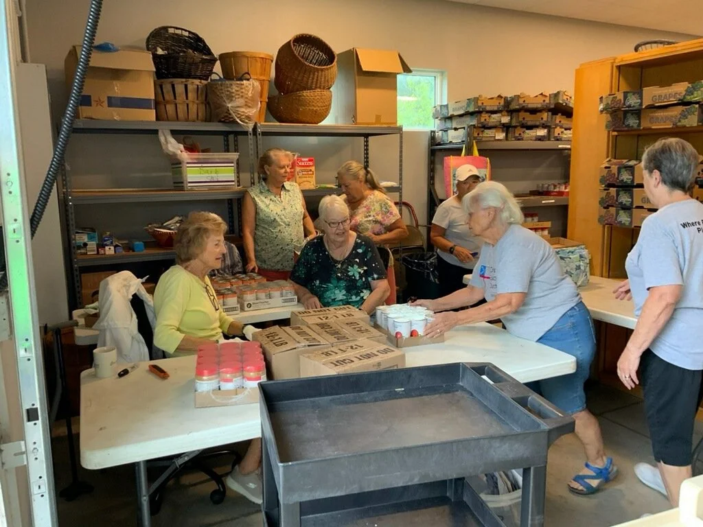 Group of people, mostly women, gathered around a table in a storage room, packing items into boxes and jars.
