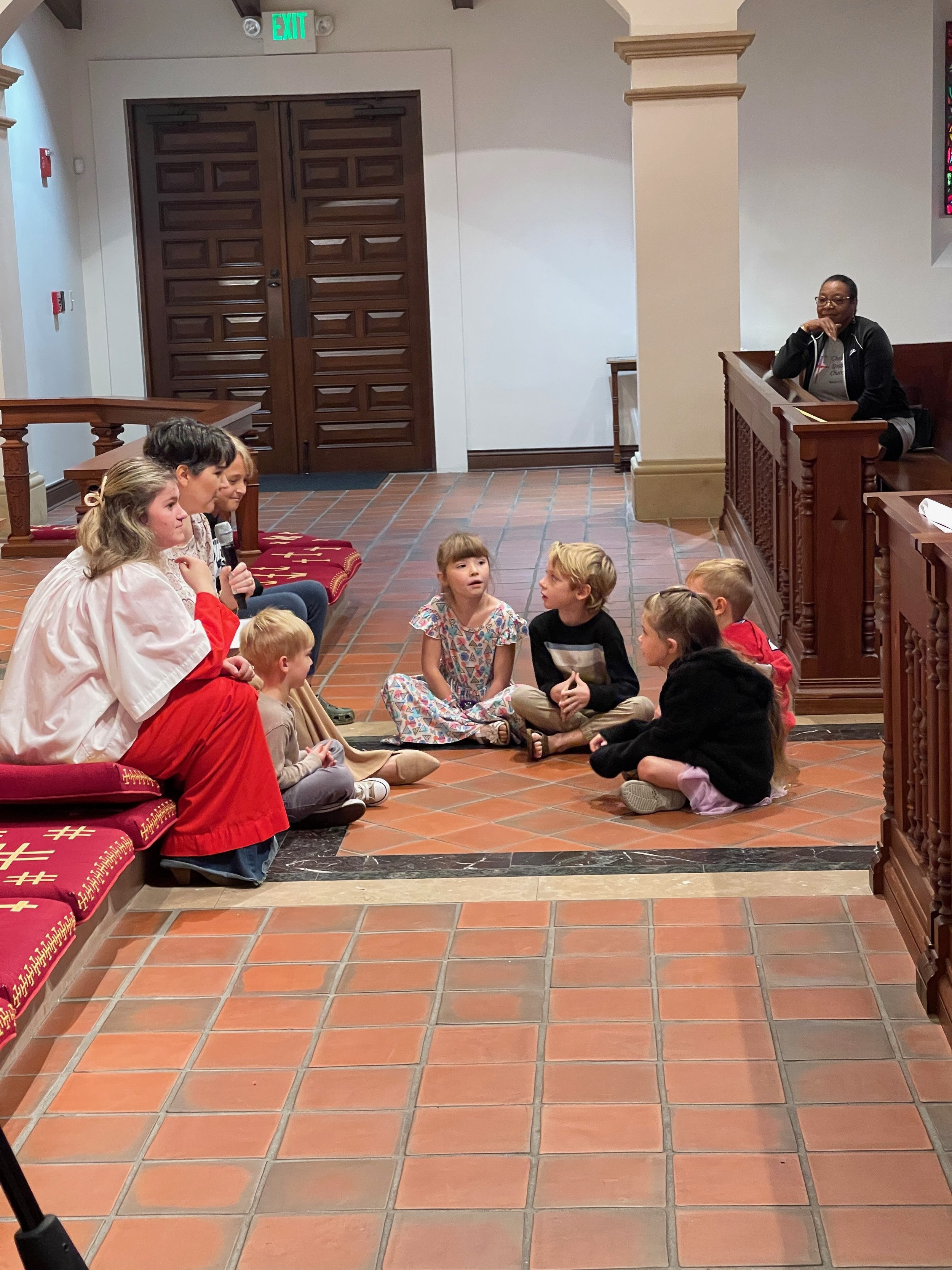 Children sitting on the floor and on benches in a church, listening to a woman with a microphone, with another woman sitting at a pulpit to the side.