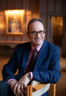 A man with glasses, wearing a dark suit and red tie, sitting and smiling indoors with warm lighting and paintings on the wall behind him.