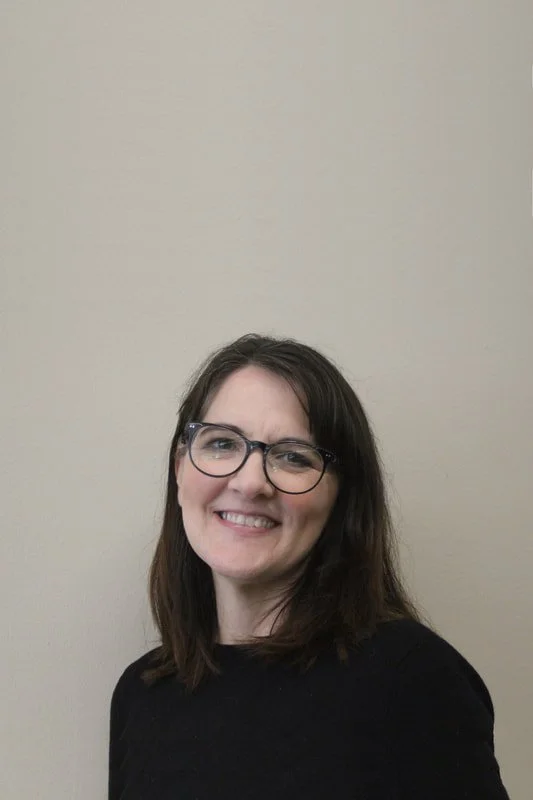 Smiling woman with glasses and dark hair, wearing a black top, standing against a plain beige wall.