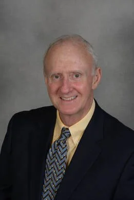 A professional headshot of an elderly man with short gray hair, wearing a black suit, yellow shirt, and patterned tie, smiling against a gray background.