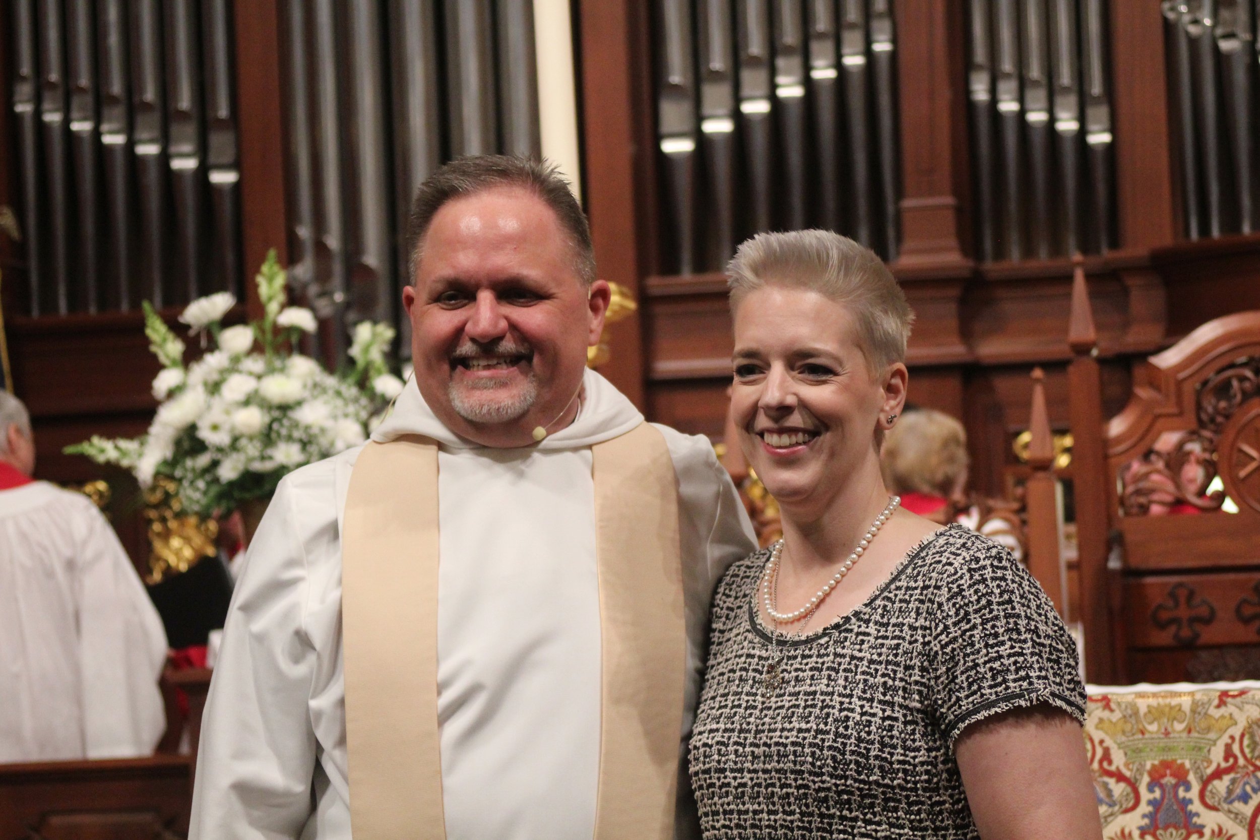 A smiling officiant and a woman standing together inside a church, with an organ and floral arrangement in the background.