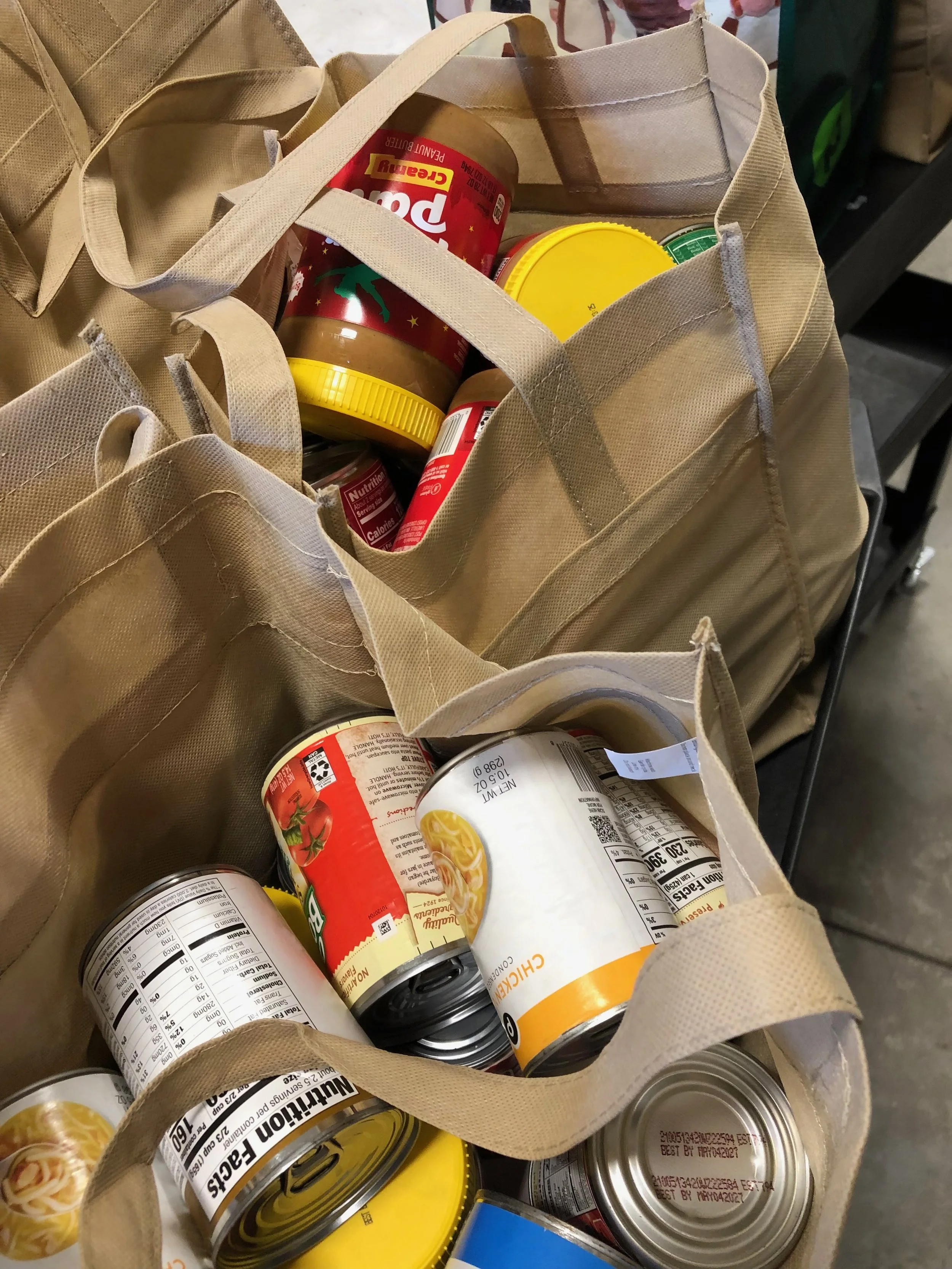 Reusable beige tote bags filled with canned foods, including chicken and tomato products, on a shopping cart.