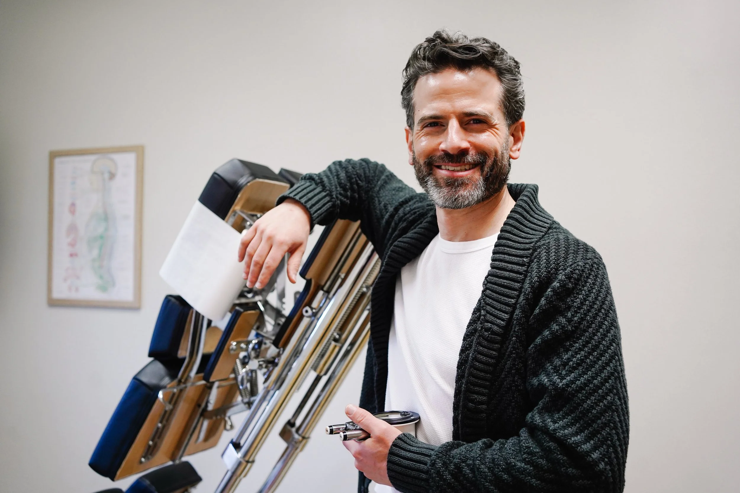 A man with dark curly hair and a beard smiles while standing in a room with medical equipment, wearing a white shirt and a black cardigan, holding a stethoscope.