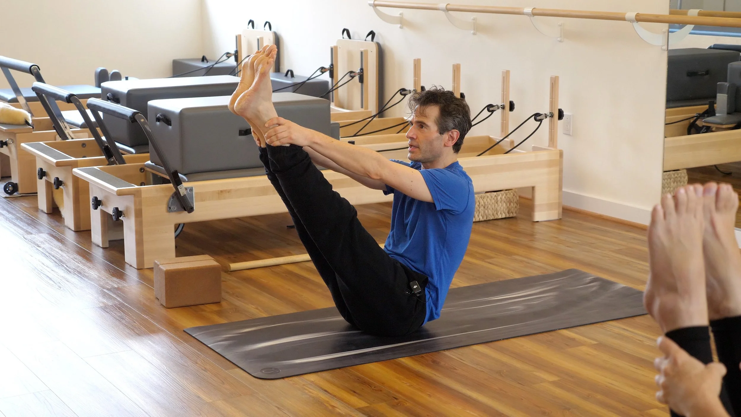 A man practicing yoga on a black mat inside a Pilates studio, sitting on the floor with legs raised and holding his feet, with Pilates equipment such as reformers and blocks in the background.