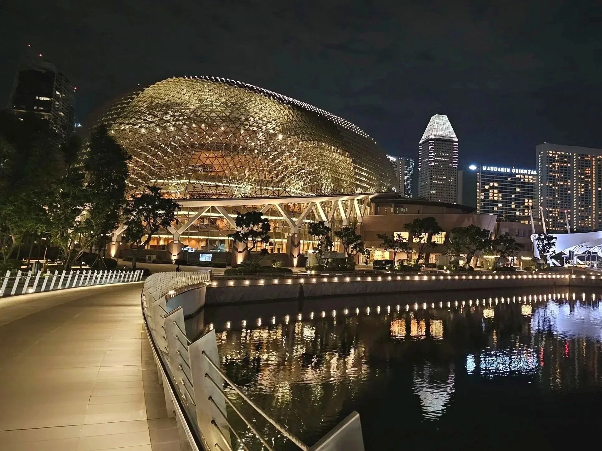 Night view of a modern building with a glass and metal dome, lit up and reflected in water, with a walking path in the foreground and other tall buildings illuminated in the background.