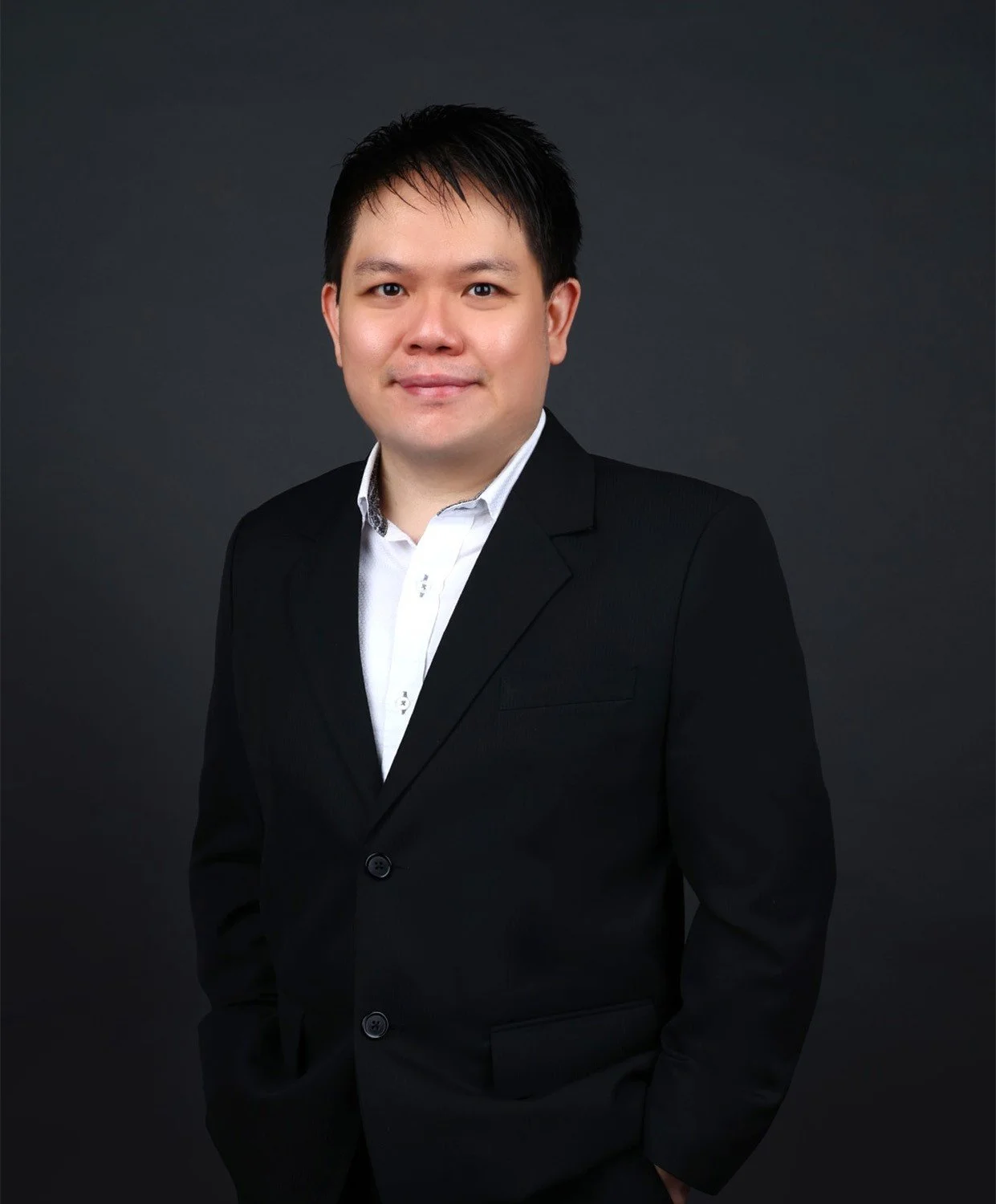Portrait of a young man with short black hair, wearing a black suit and white shirt, standing against a dark gray background.