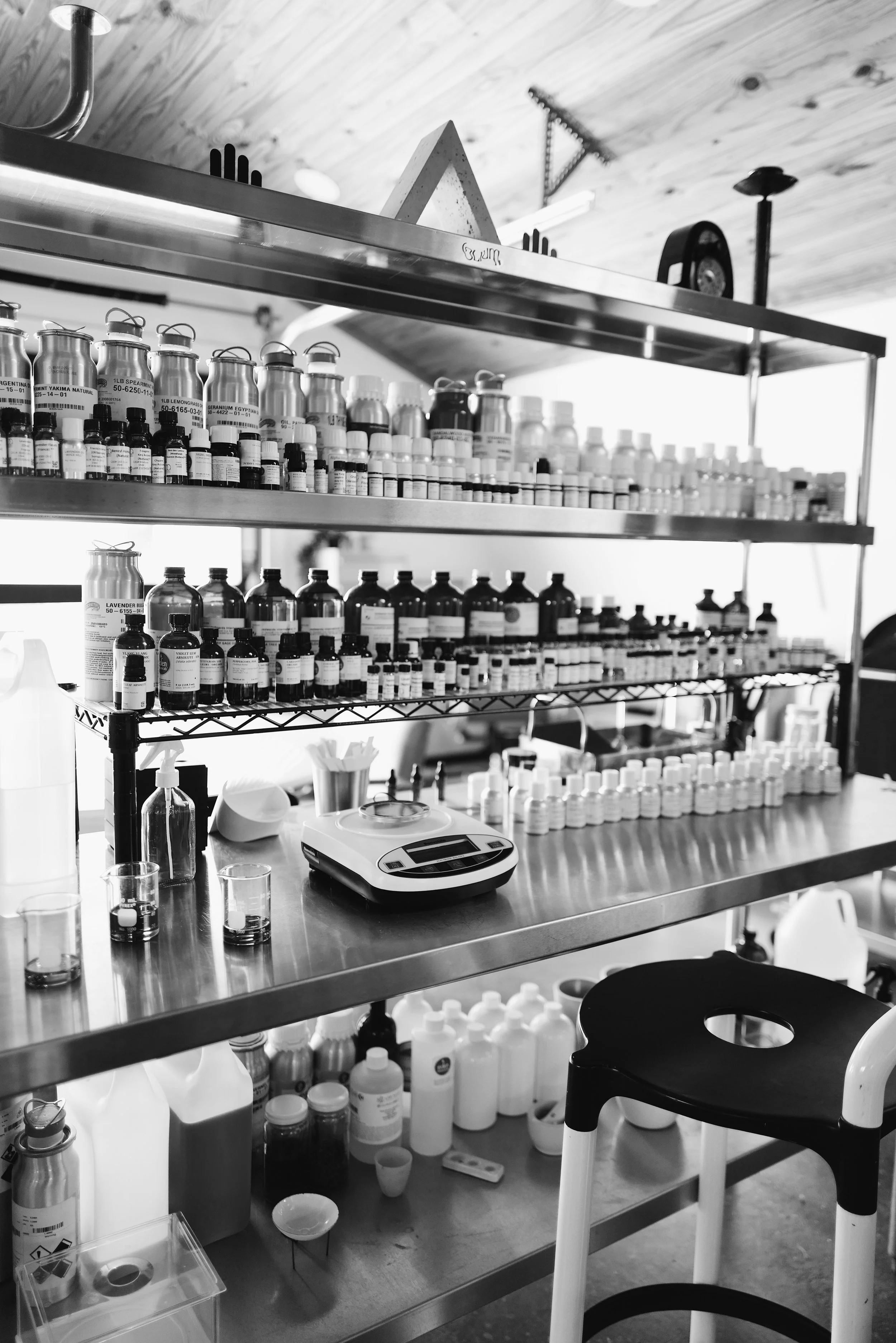A metal shelving unit holding small bottles and jars, a digital scale, and various containers, with a designer Kartell stool nearby in a perfumery science lab setting.