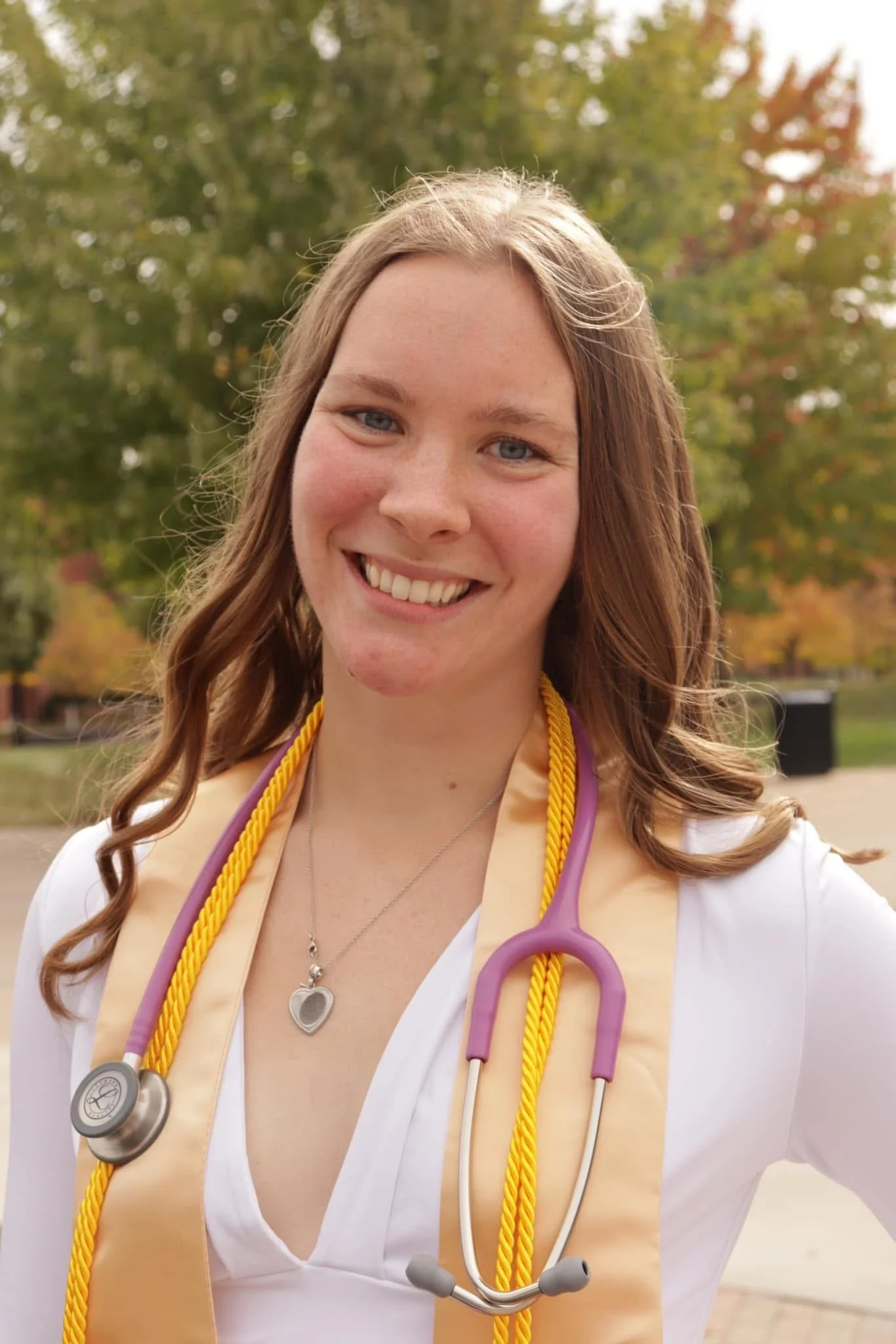 A smiling young woman (Lily) with long, wavy brown hair, wearing a white top, a yellow graduation stole, and a stethoscope around her neck, standing outdoors in a park with trees in fall colors.