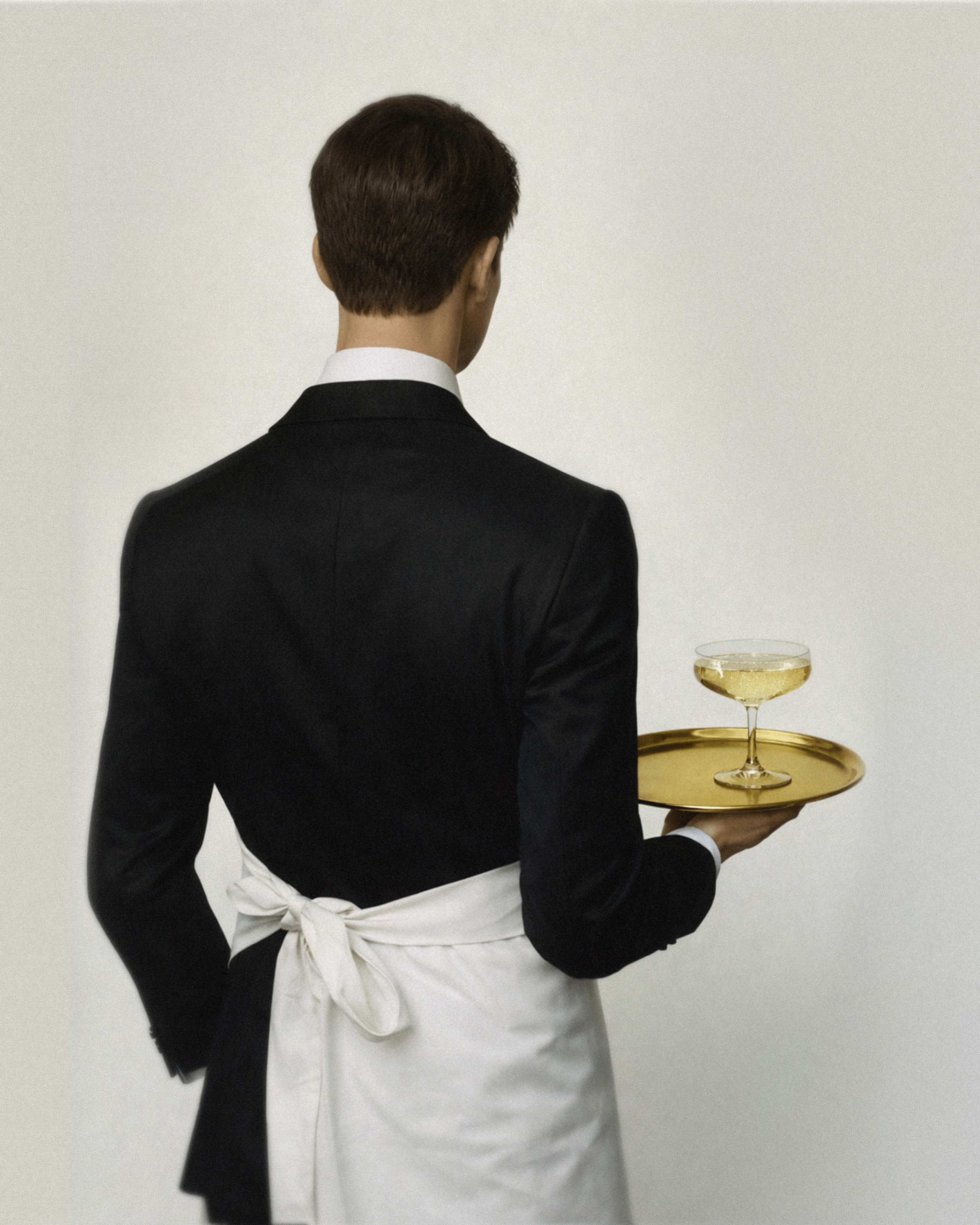Back view of a waiter in a black suit and white apron carrying a gold tray with a coupe glass of champagne.