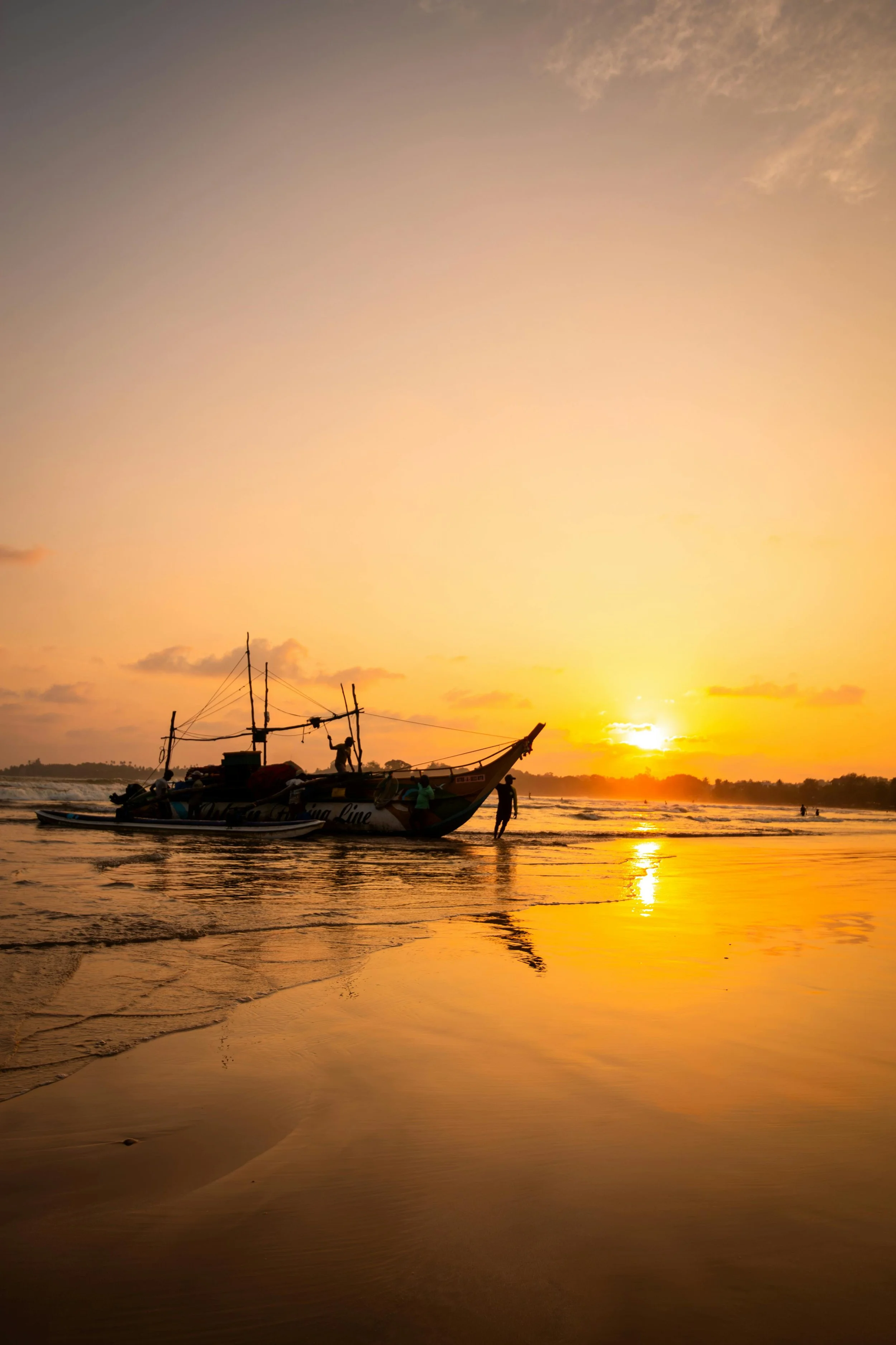 A boat with multiple people on it is anchored near the shoreline during sunset, with the sky painted in warm hues of orange and pink, and the water reflecting the colors of the sky.