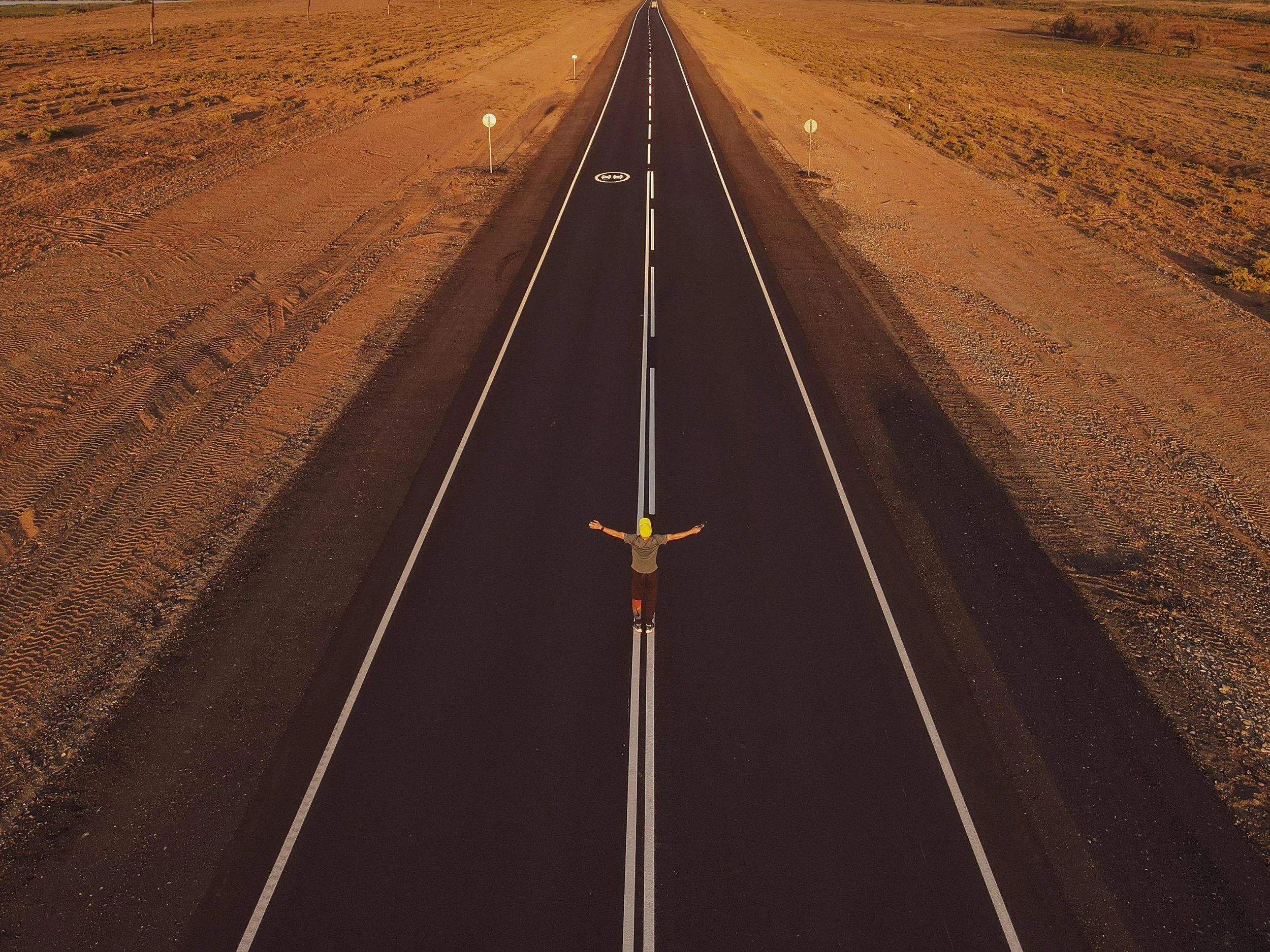 A person wearing a yellow cap, gray shirt, and dark pants standing in the middle of a long empty paved road extending into the horizon. The person has arms outstretched and appears to be enjoying the open road. Surrounding the road is a barren, desert-like landscape with reddish-brown soil and little vegetation, under a clear sky.