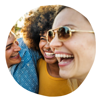 Three diverse friends smiling and laughing together outdoors, with sunlight and blurred trees in the background.
