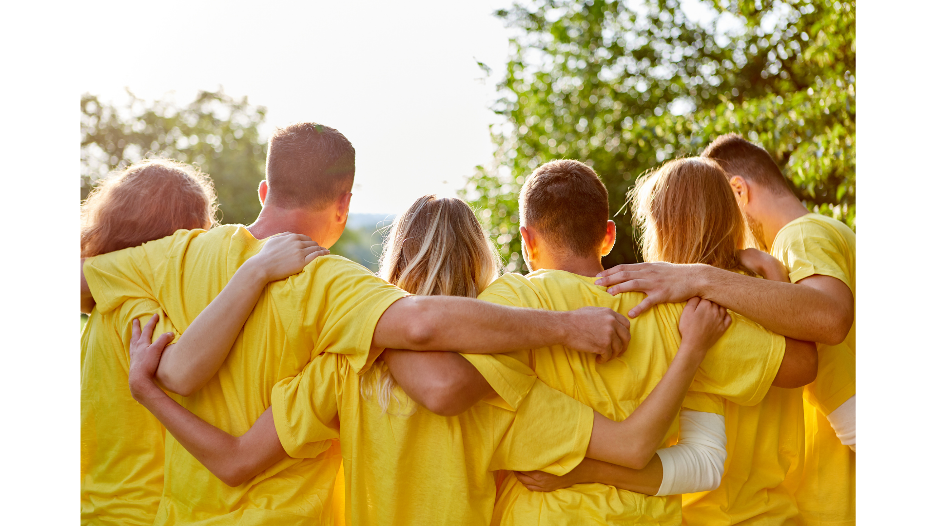 A group of people wearing yellow shirts standing closely together outdoors, embracing with their arms around each other, facing away from the camera.