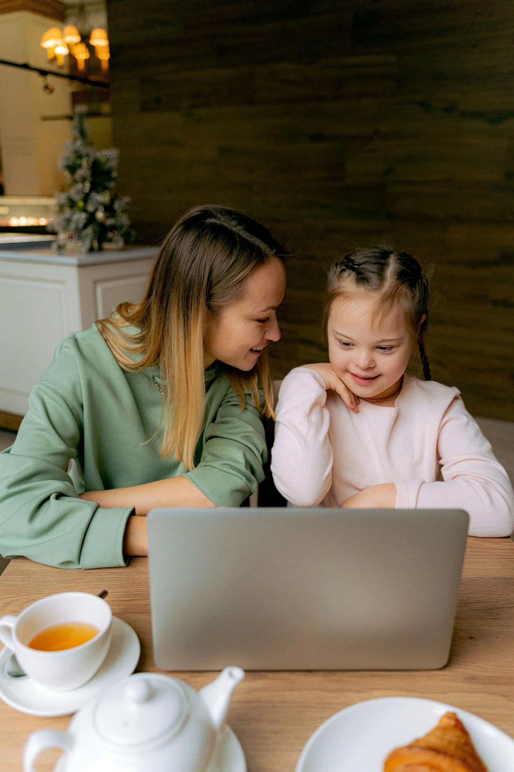 A woman and a girl looking at a laptop screen together at a table, with a cup of tea and a croissant in the foreground.