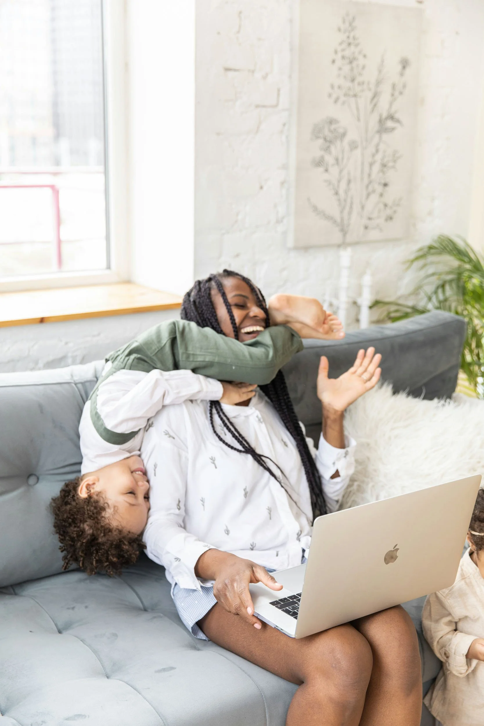 A woman sitting on a gray couch with a child leaning on her, using a laptop, laughing and playing together in a bright living room.