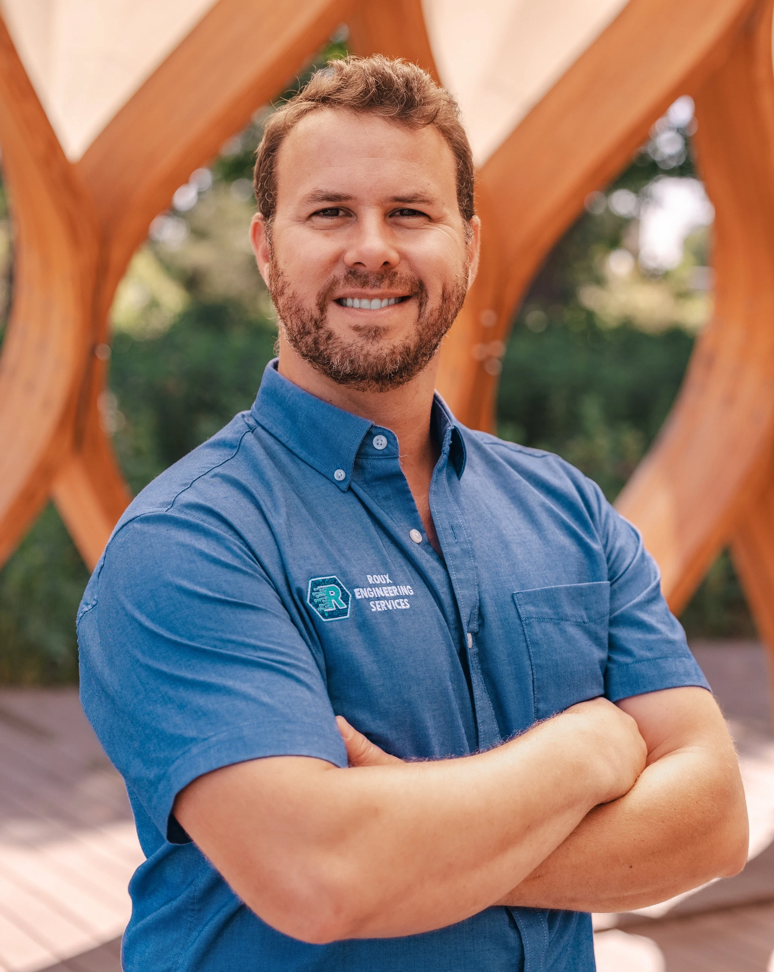 A man with a beard and short brown hair standing outdoors with his arms crossed, smiling, wearing a blue shirt with a logo that reads 'Roux Engineering Services,' and a wooden structure in the background.