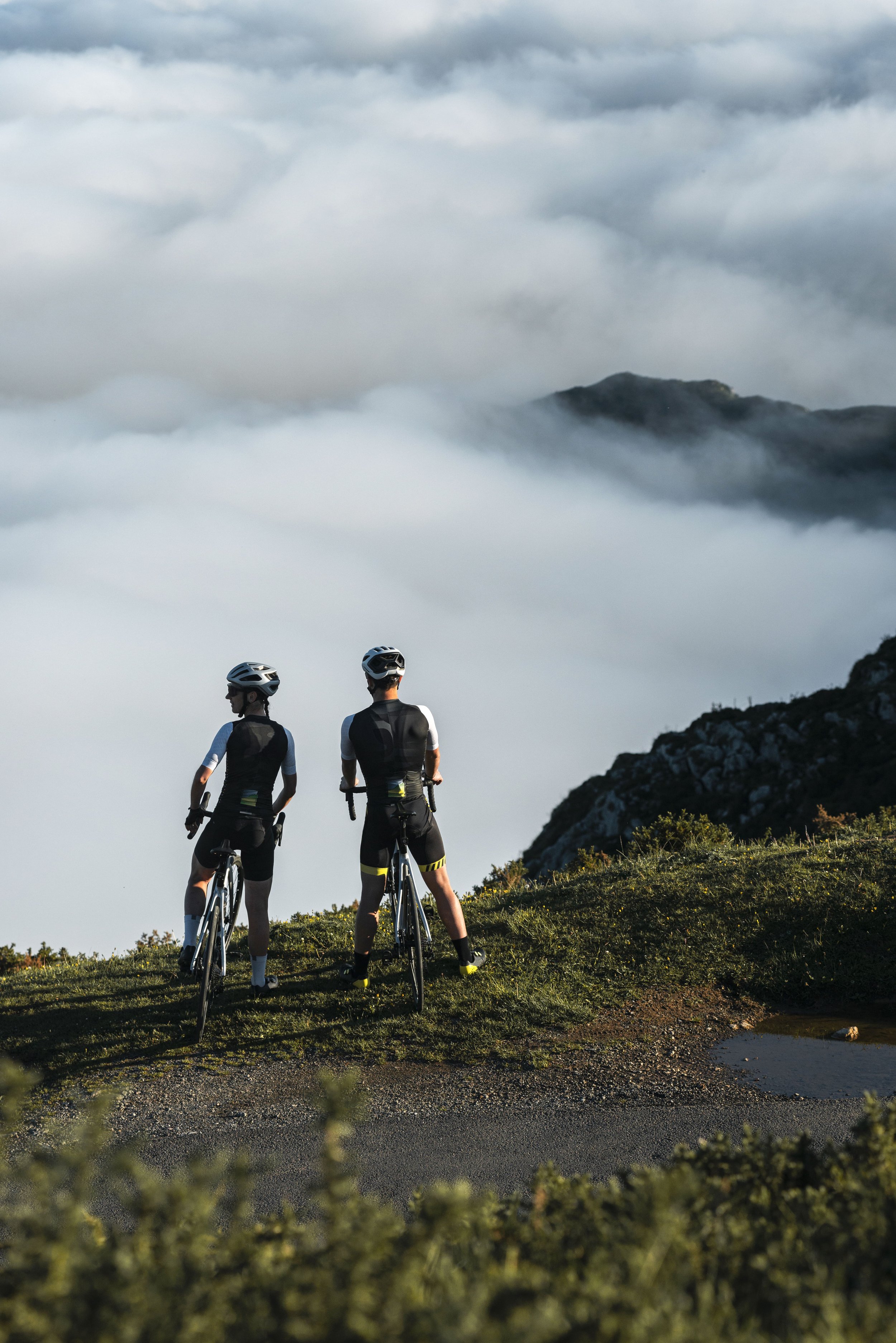 Amigos entrenando juntos en bicicleta por una carretera rural, rodeados de montañas cubiertas de niebla durante el verano