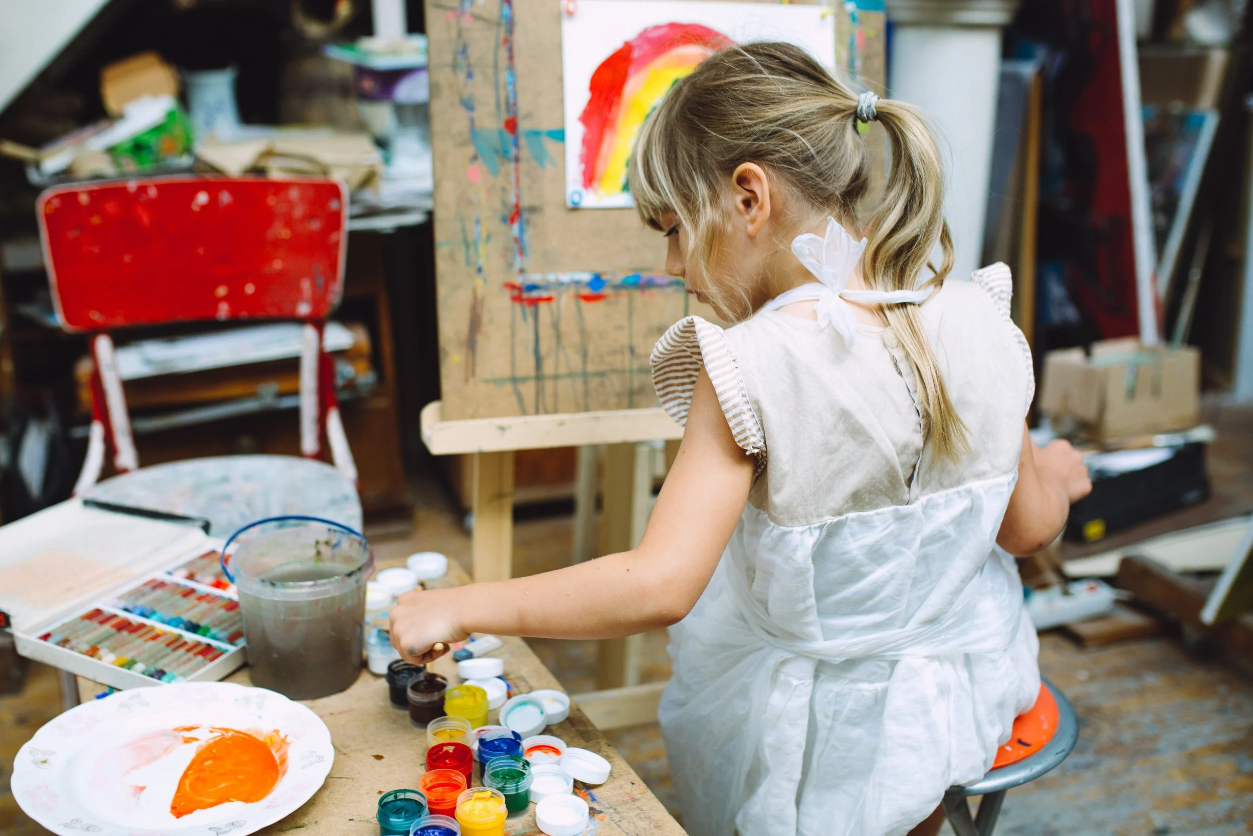 A young girl with a ponytail wearing a white apron painting a colorful rainbow on a paper at an art studio, surrounded by paints, brushes, and art supplies.