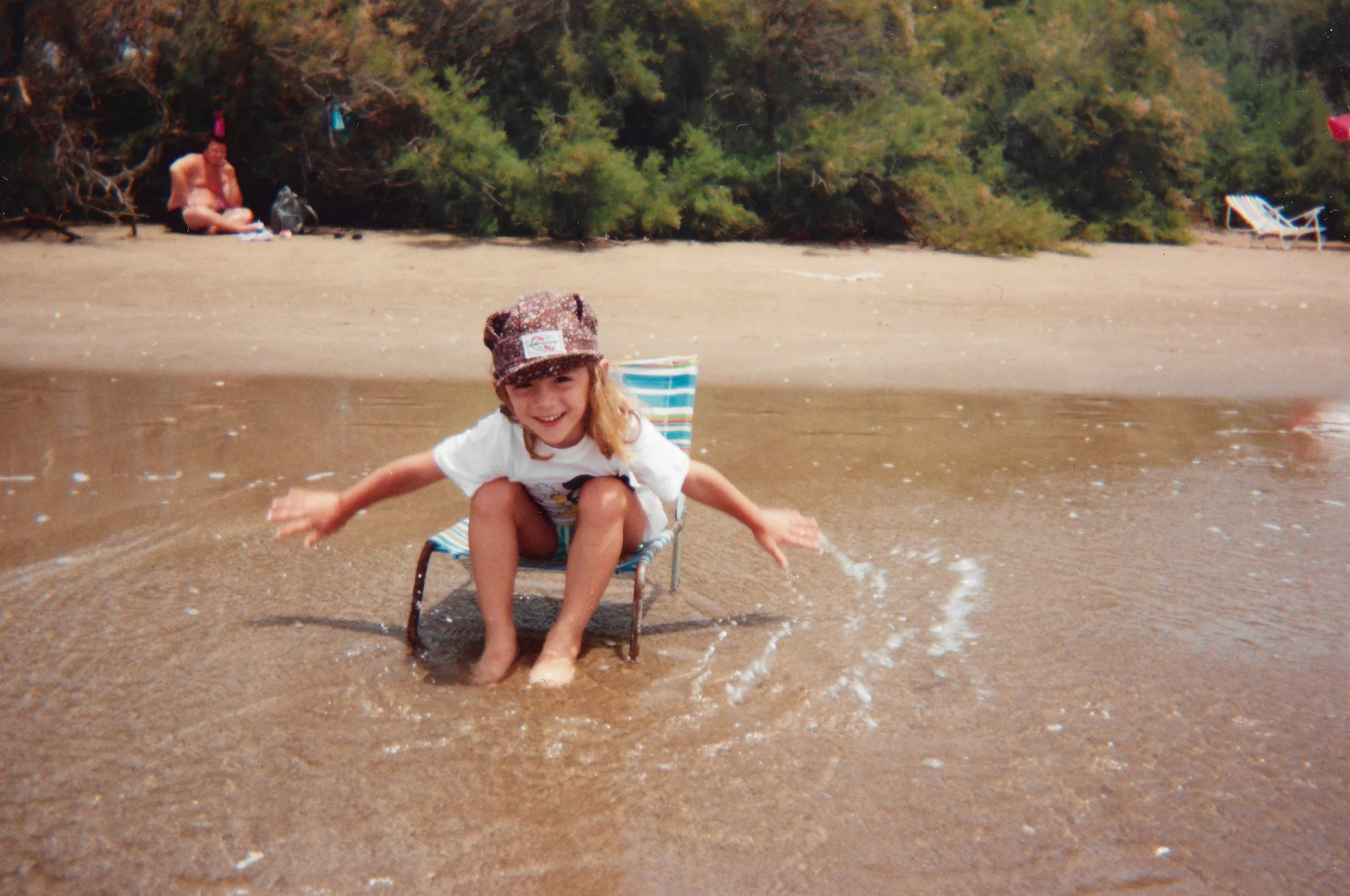 Young girl with a hat smiling on a beach chair in shallow water during daytime with trees and a woman sitting on the sand in the background.