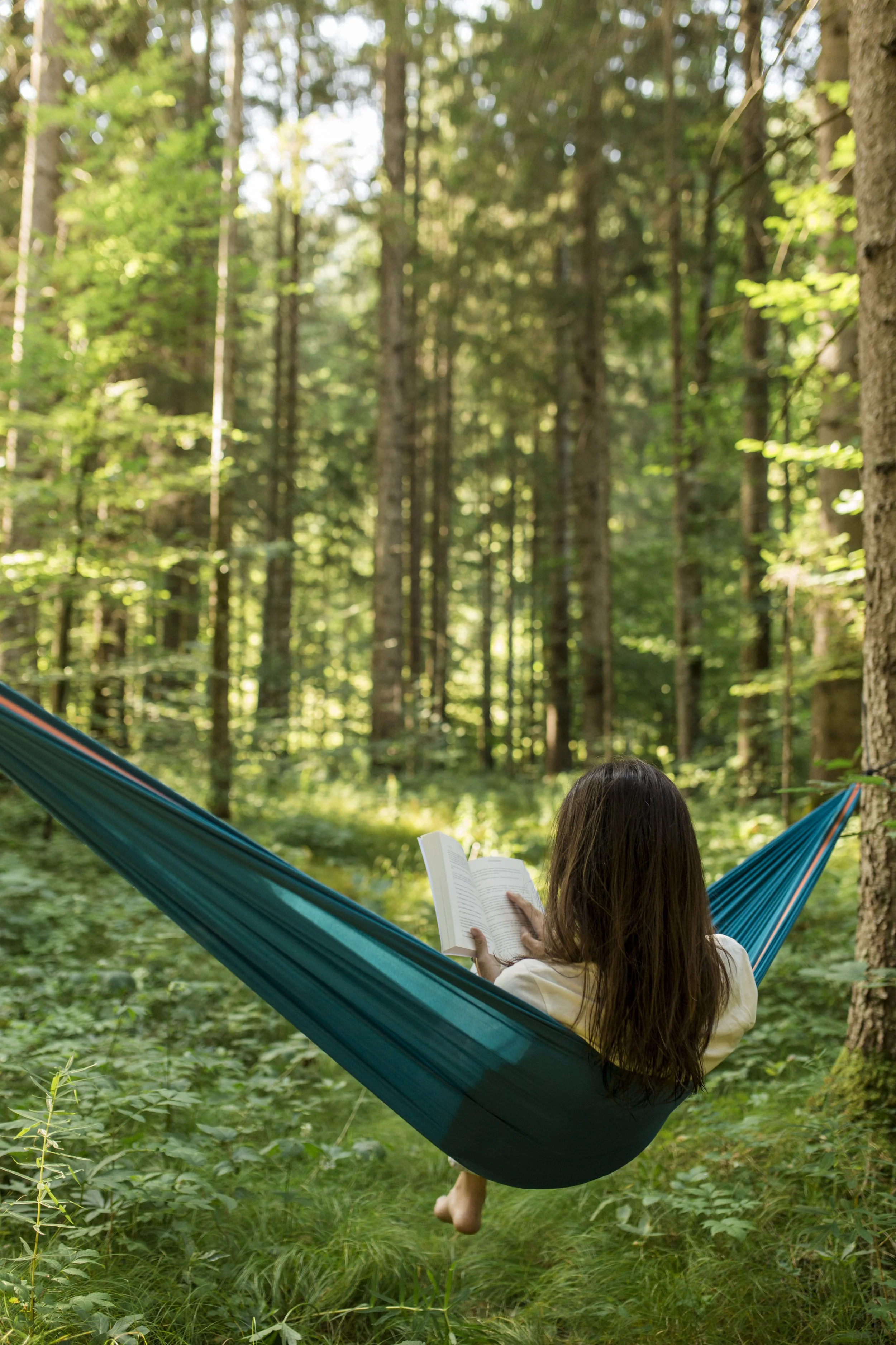 A woman with long brown hair sitting in a teal hammock, reading a book, in a lush green forest with tall trees and dense foliage.