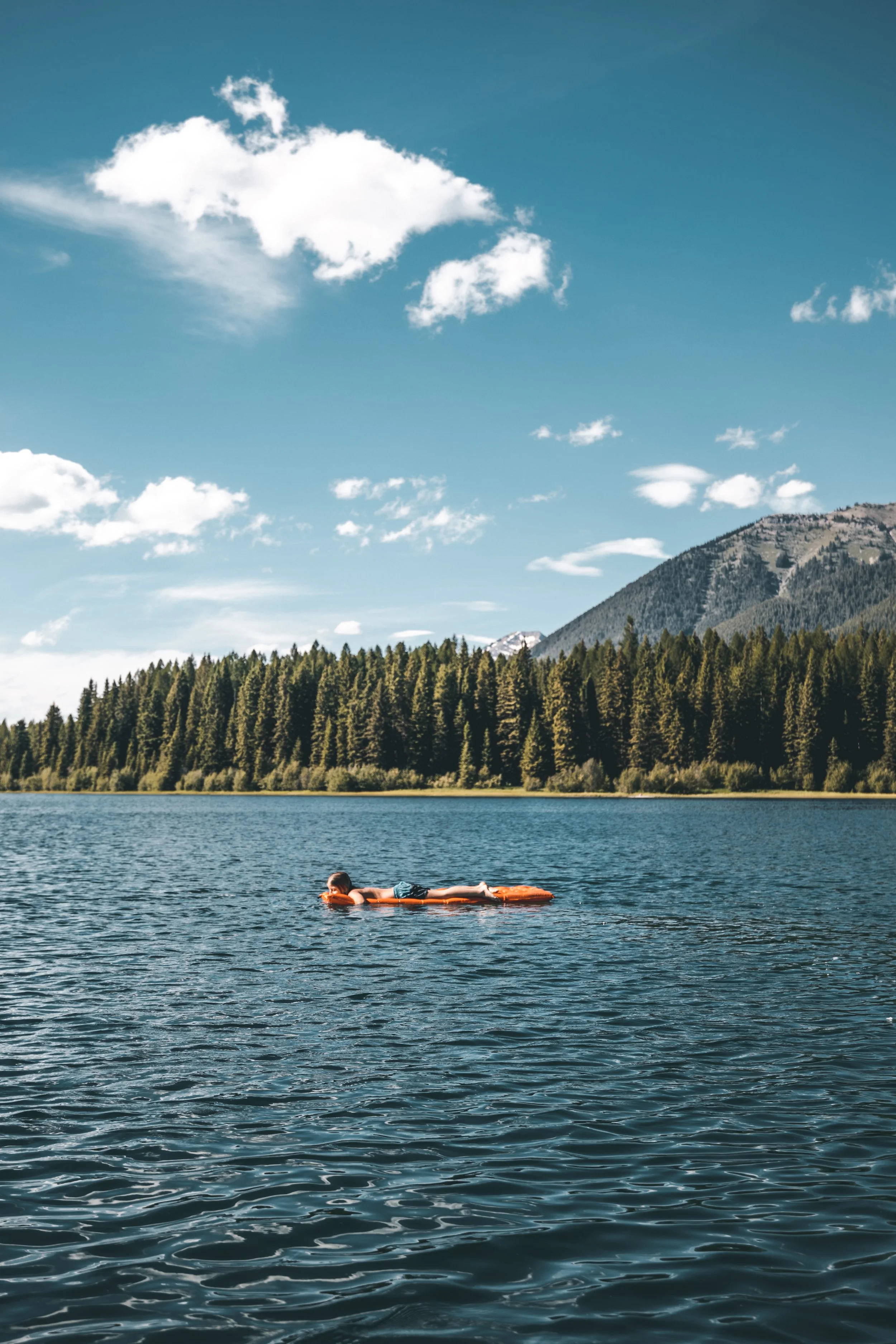 A person lying on a float in a lake with mountains and pine trees in the background under a partly cloudy sky.
