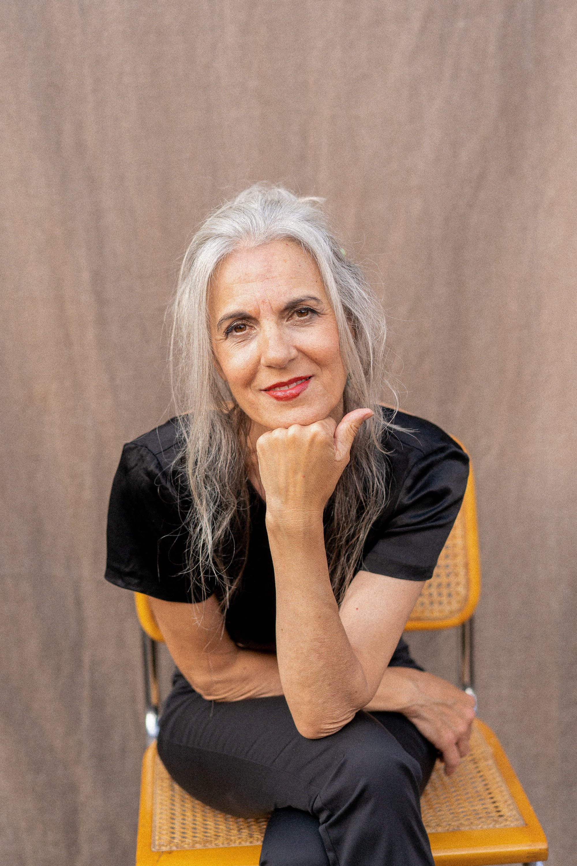 Mujer madura, segura y hermosa posando delante de un fondo. Tiene el pelo largo y gris, está maquillada y lleva un traje negro