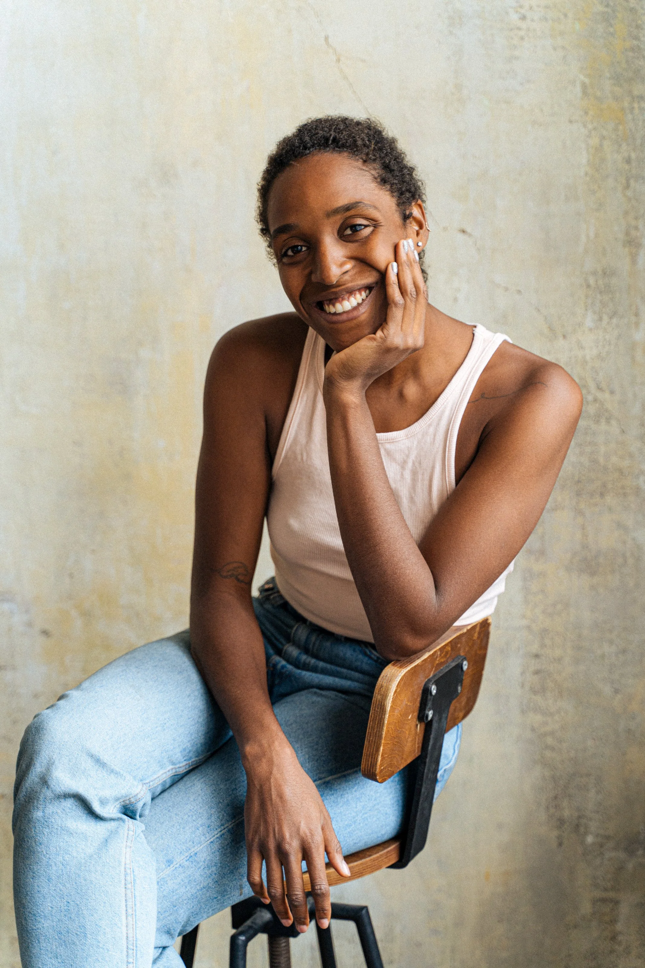 A young Black woman with short curly hair, wearing a light-colored tank top and jeans, sitting on a wooden and metal chair with her right hand touching her face, smiling with visible teeth, against a textured wall background.