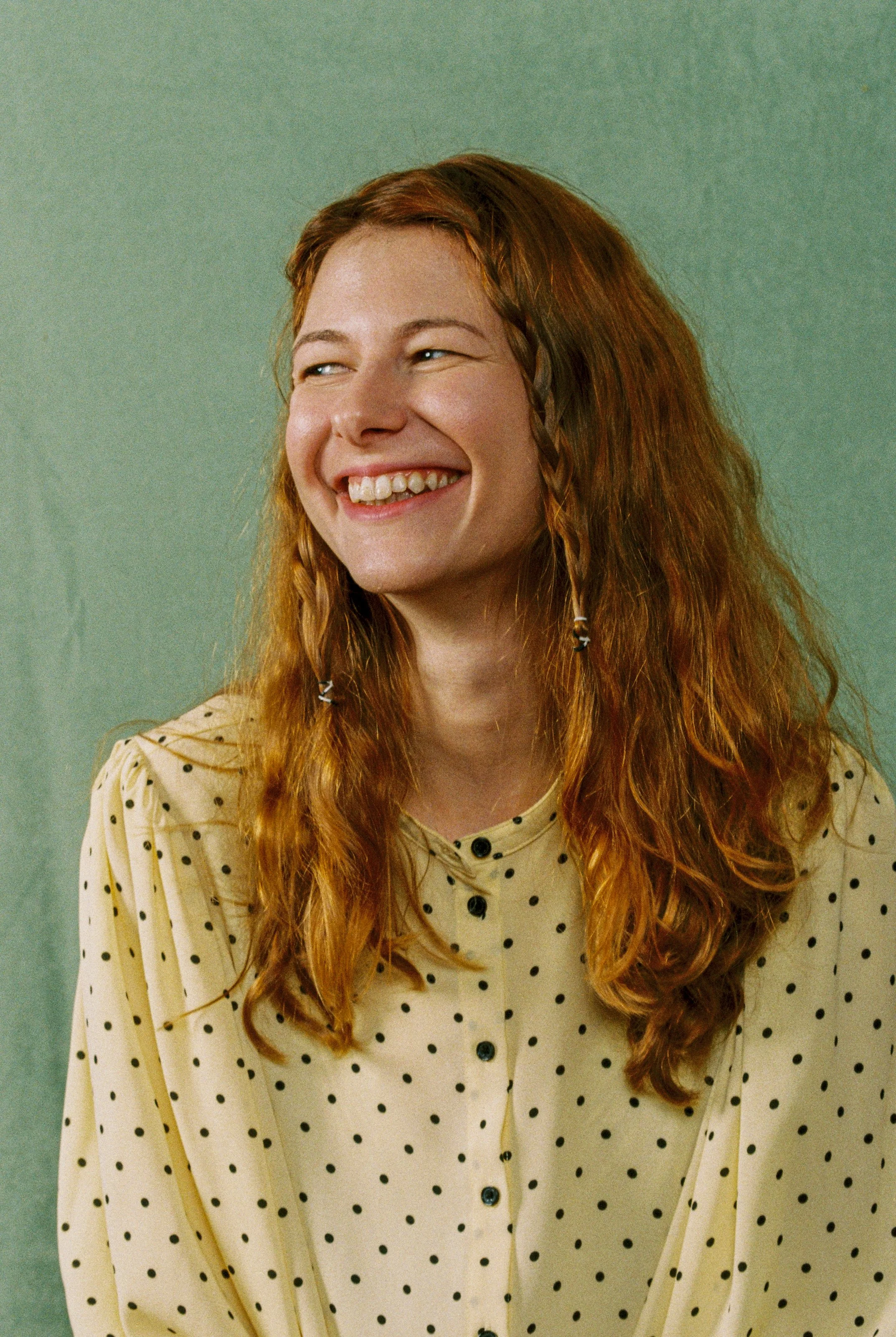 A young woman with long, curly red hair and light skin, smiling with her teeth showing. She is wearing a yellow polka dot blouse with black buttons and is against a plain green background.