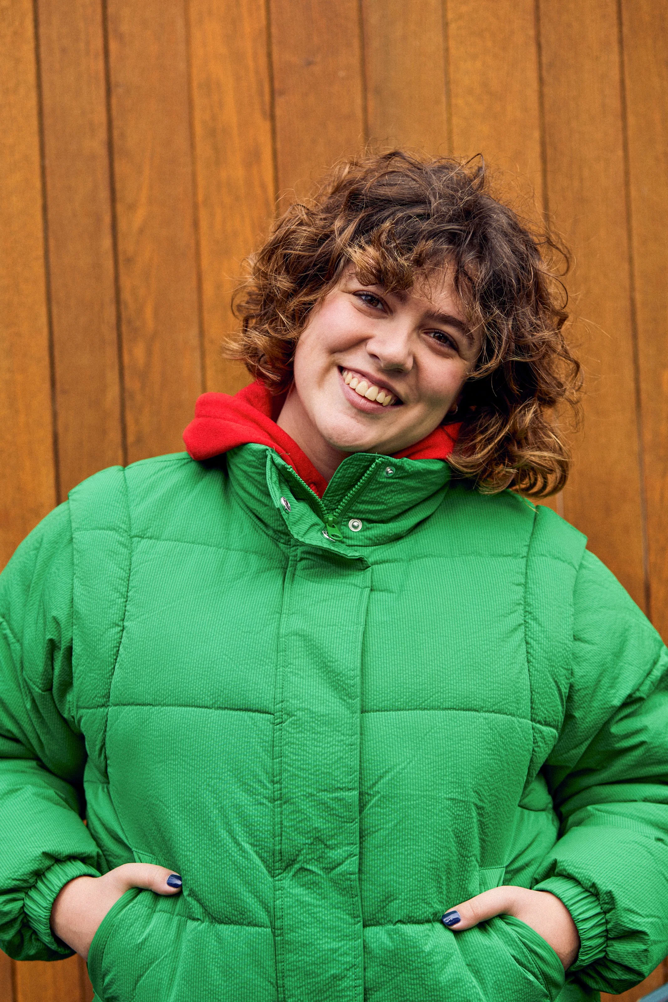 A woman with short, curly brown hair, smiling, wearing a bright green puffer jacket with a red hood layered underneath, standing against a wooden background.