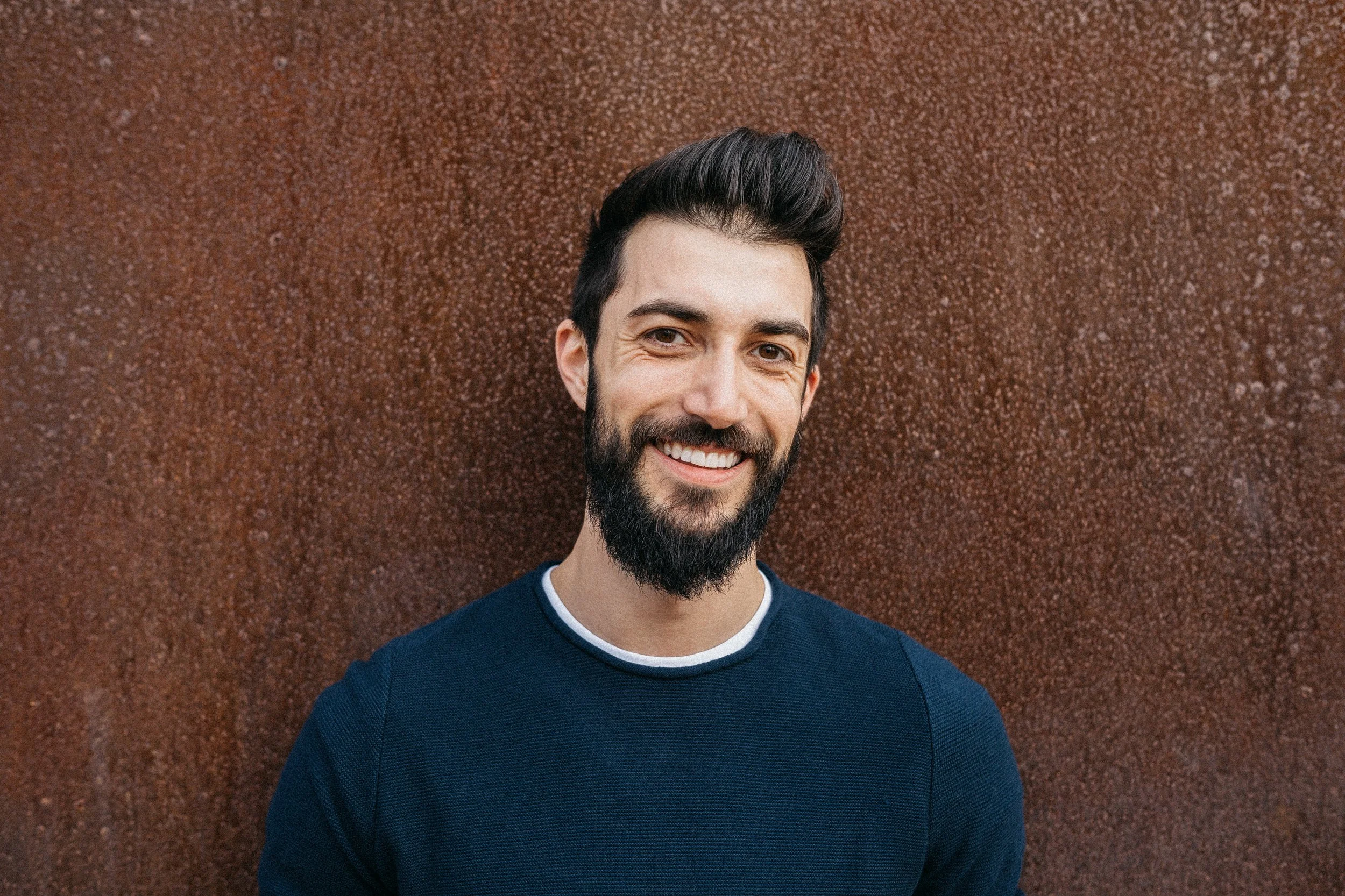 A smiling man with dark hair and a beard wearing a navy blue sweater, standing against a rust-colored textured wall.