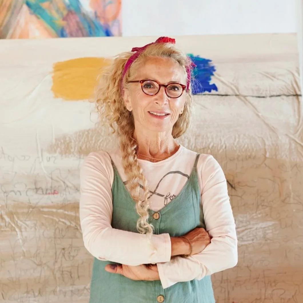 A woman with curly blonde hair, wearing glasses with red frames, a pink headband, a cream long-sleeve shirt, and a green buttoned dress. She is smiling and standing with her arms crossed in front of an abstract colorful background.
