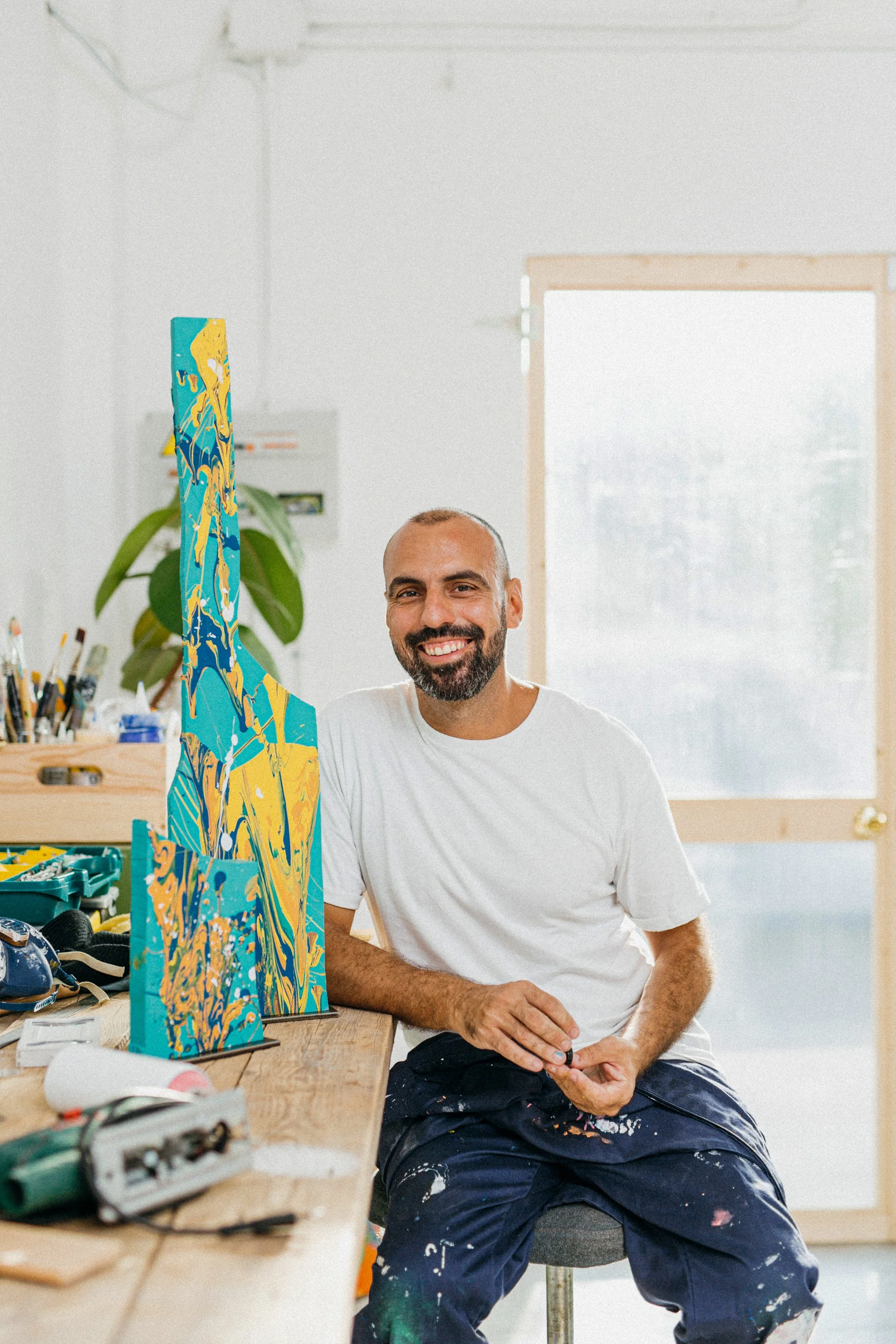 A man with a beard and a smile, sitting at a workbench in a bright art studio, smiling at the camera. The workbench has a colorful abstract painting on a canvas, and there are art supplies such as brushes and tools around.