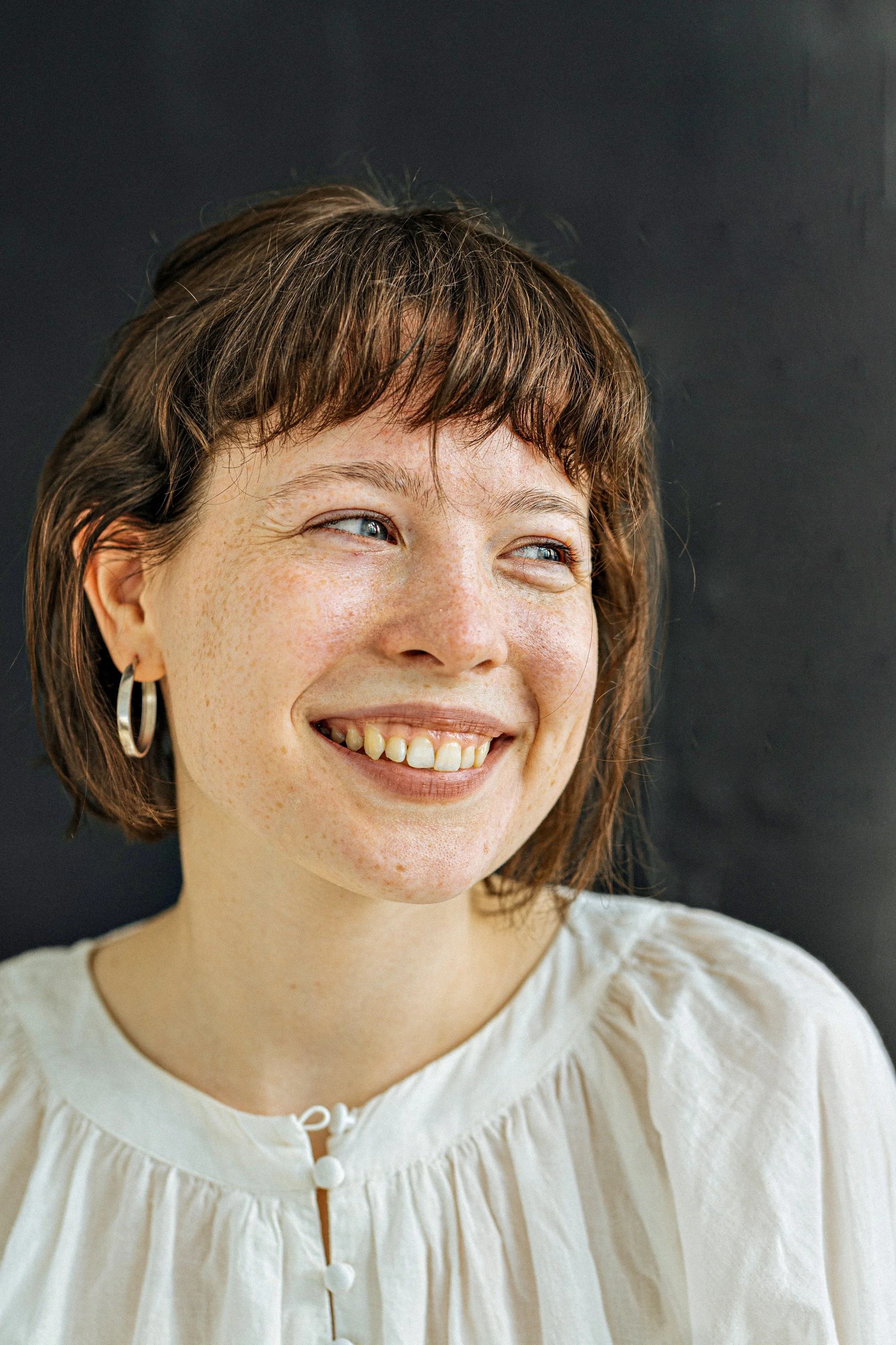 Close-up of a smiling woman with short brown hair, blue eyes, freckles, wearing silver hoop earrings, and a cream-colored top, against a dark background.