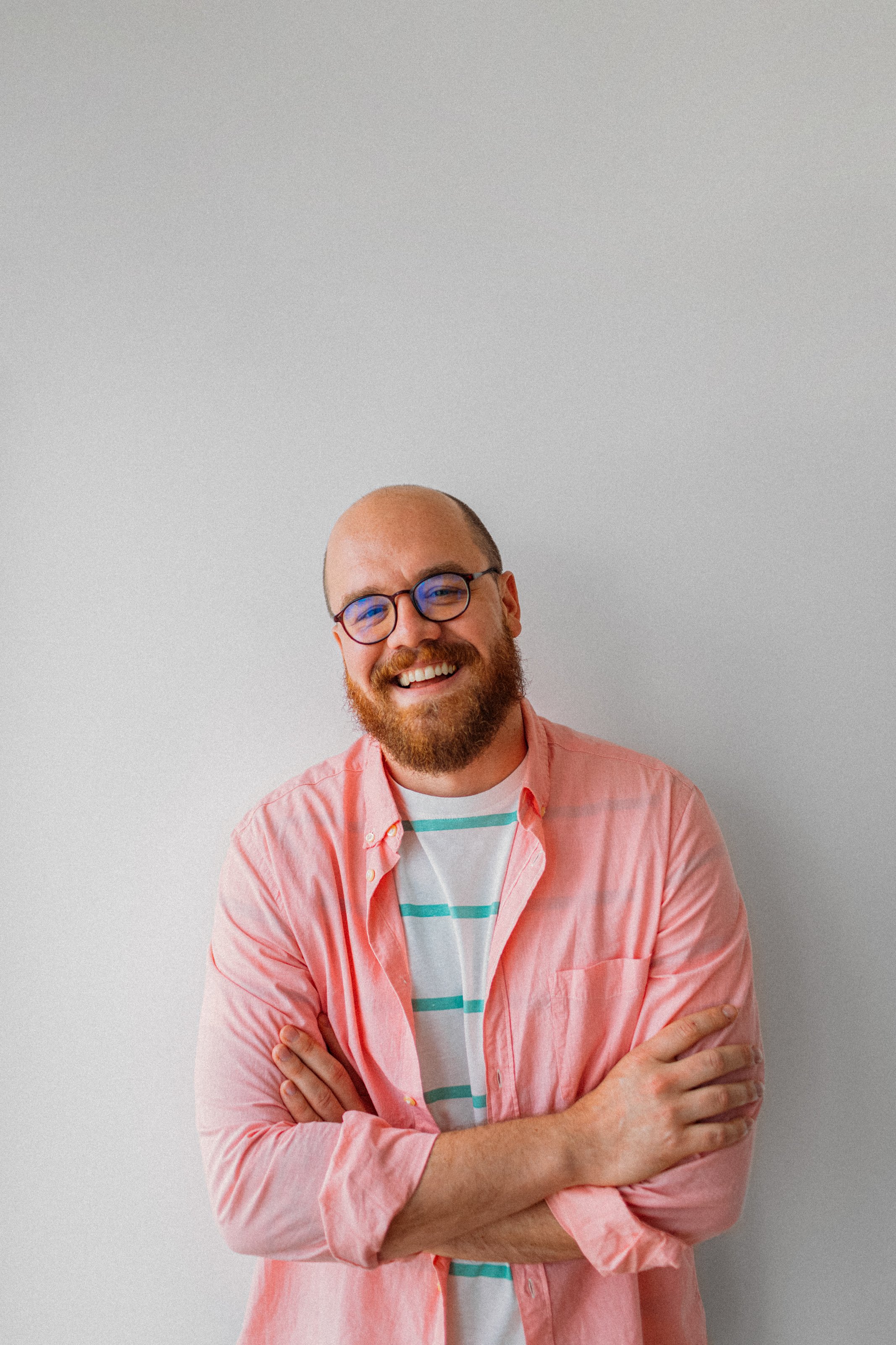 A man with glasses and a beard smiling, wearing a pink button-up shirt over a striped t-shirt, standing against a plain light gray wall with arms crossed.