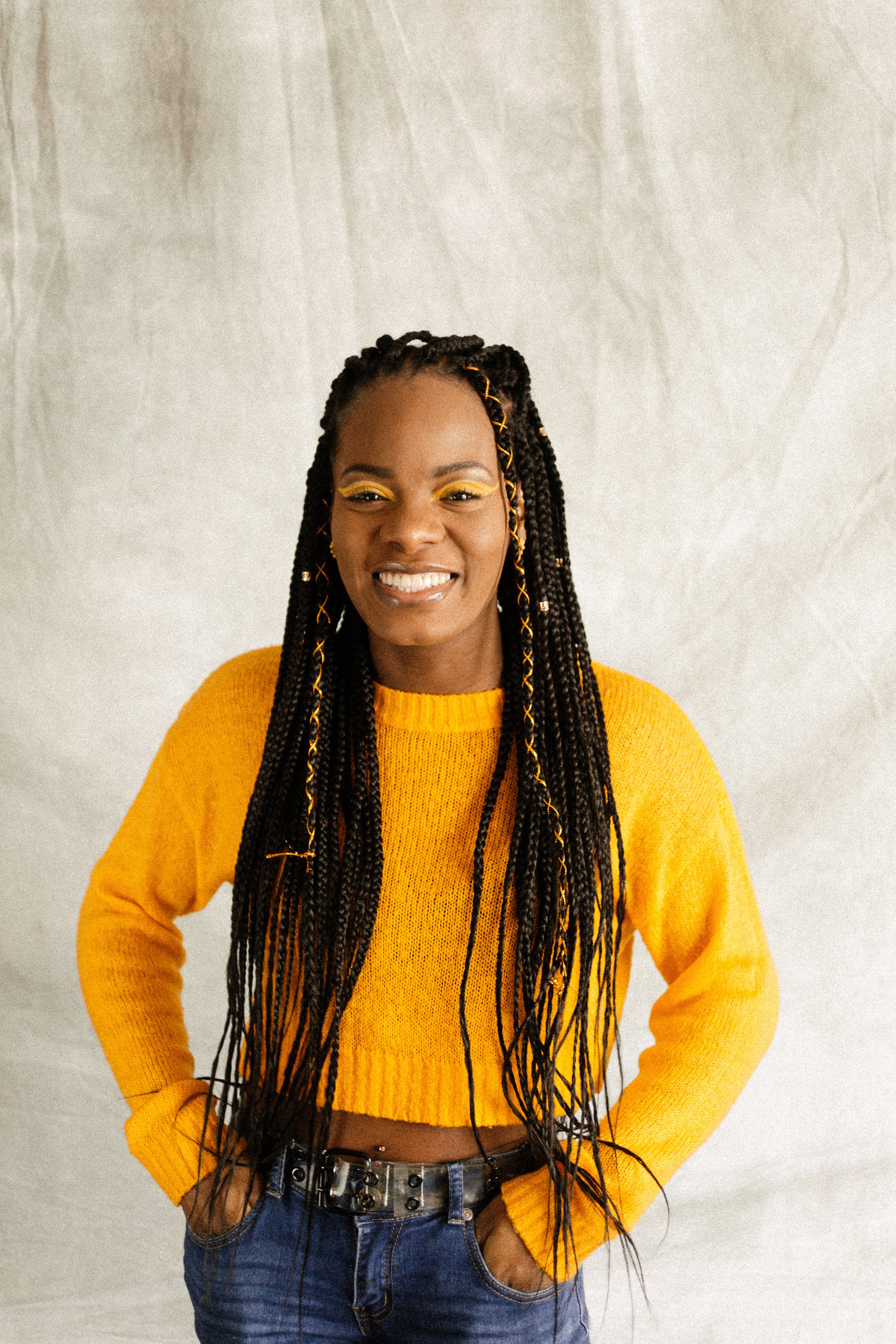A woman with long braids wearing a yellow sweater and blue jeans, smiling, standing against a plain, light-colored background.