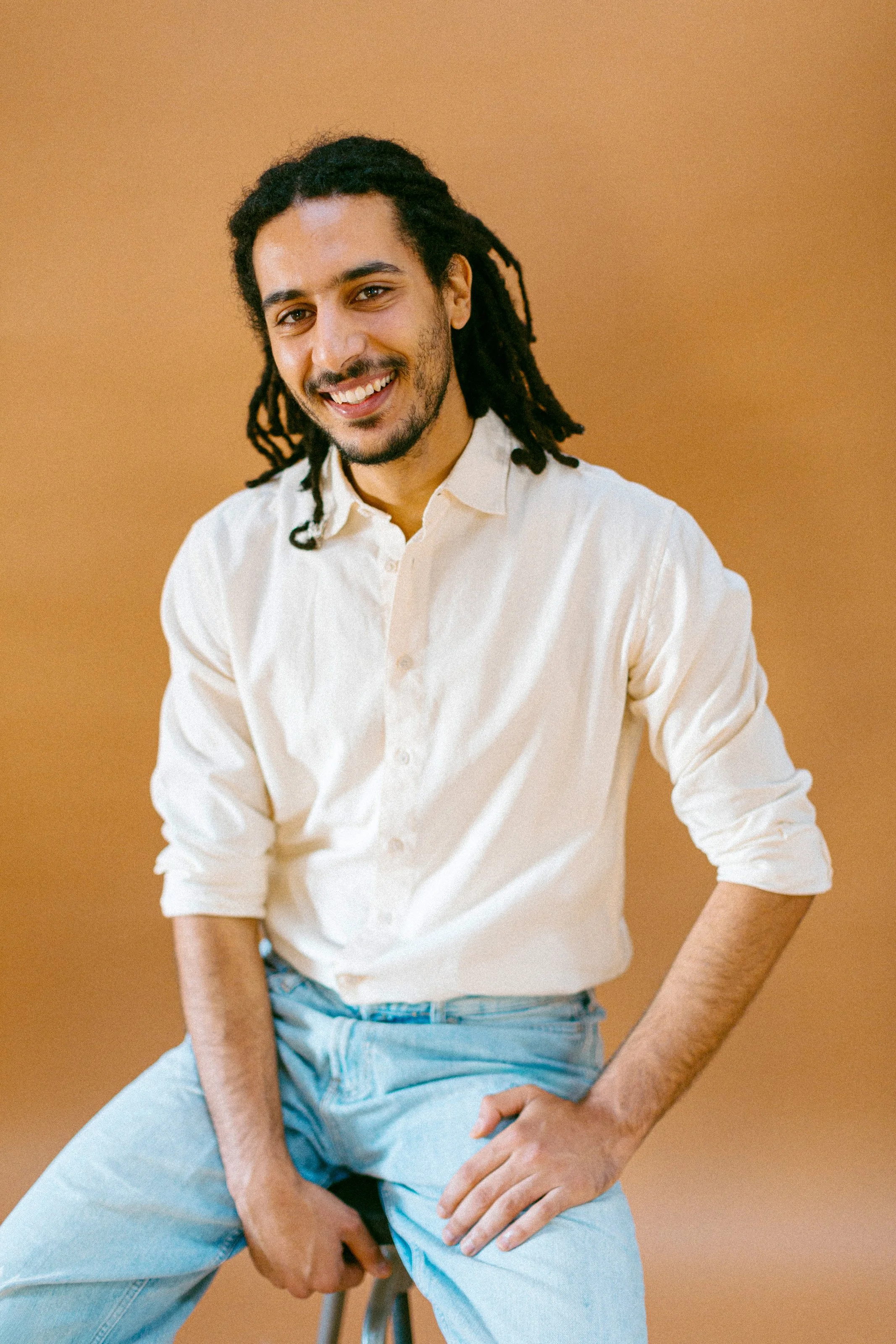 A young man with dreadlocks and a beard, wearing a white button-up shirt with the sleeves rolled up and light blue jeans, sitting on a stool against a tan background, smiling at the camera.