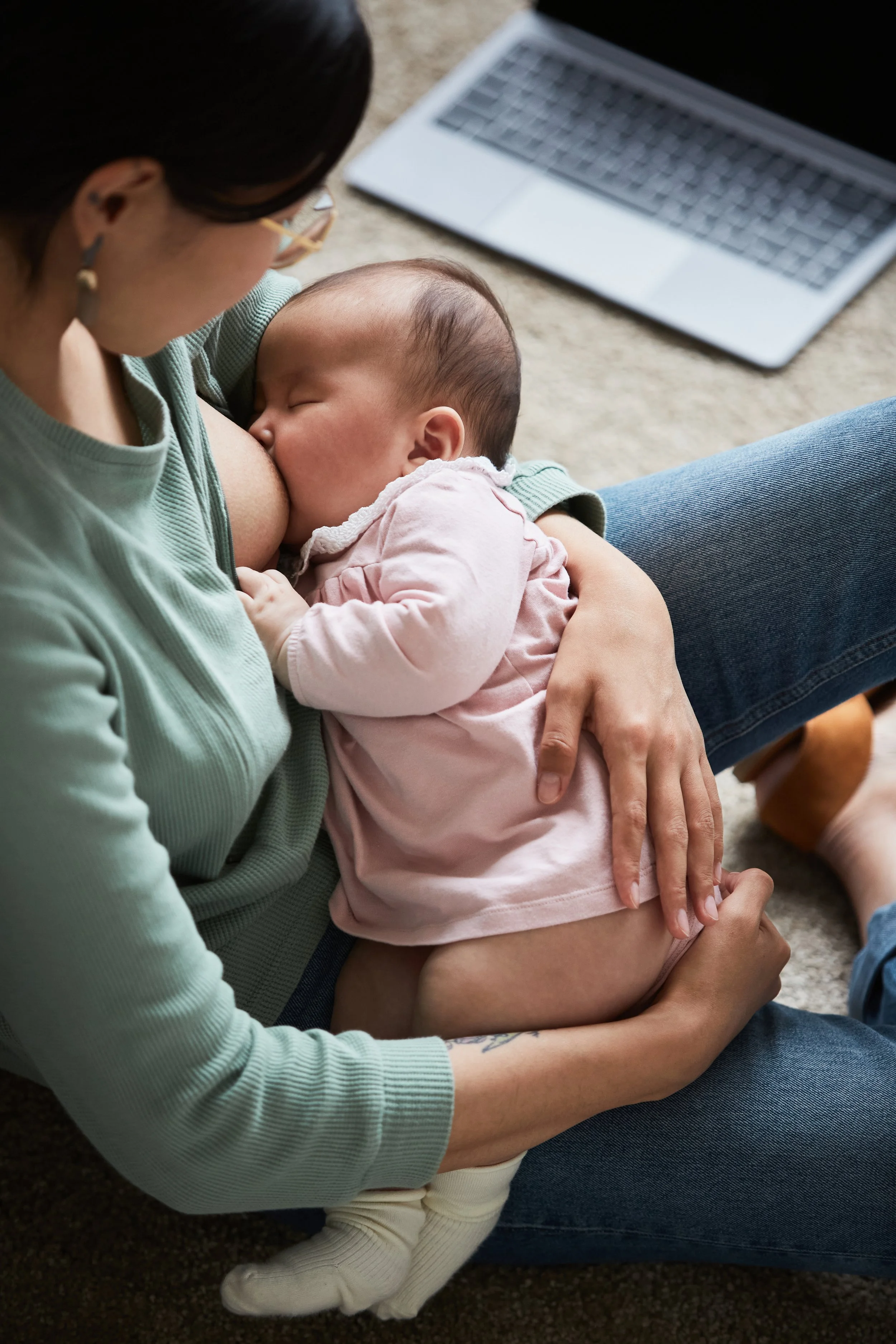 A woman with dark hair, glasses, and earrings is holding a young child. The child, with short hair, is nursing while sitting on her lap. A laptop is on the floor nearby, and the scene takes place on a carpeted surface.