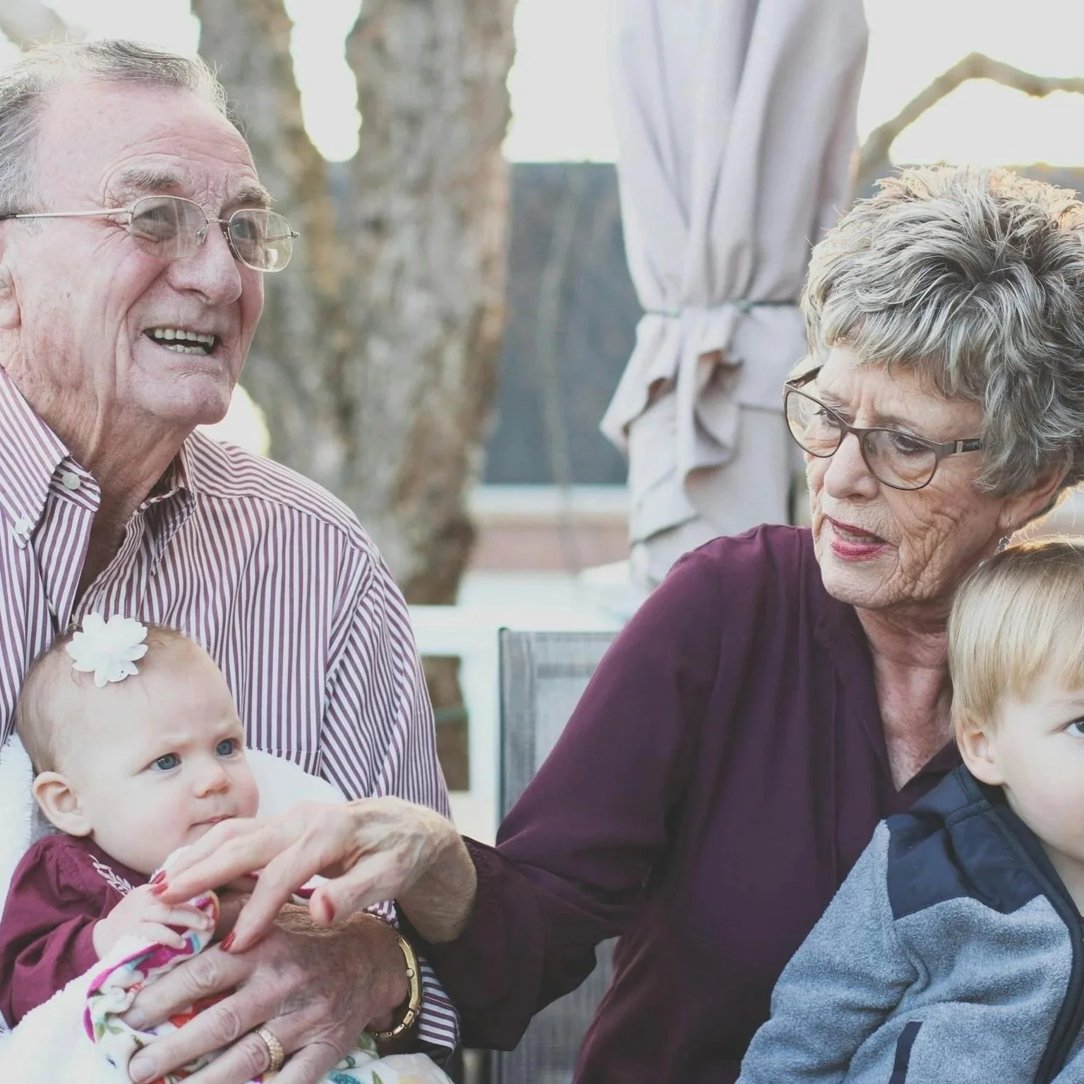 Older man with glasses and striped shirt holding a baby girl with a white flower headband, sitting outdoors with an elderly woman with glasses and gray hair, and a young boy.