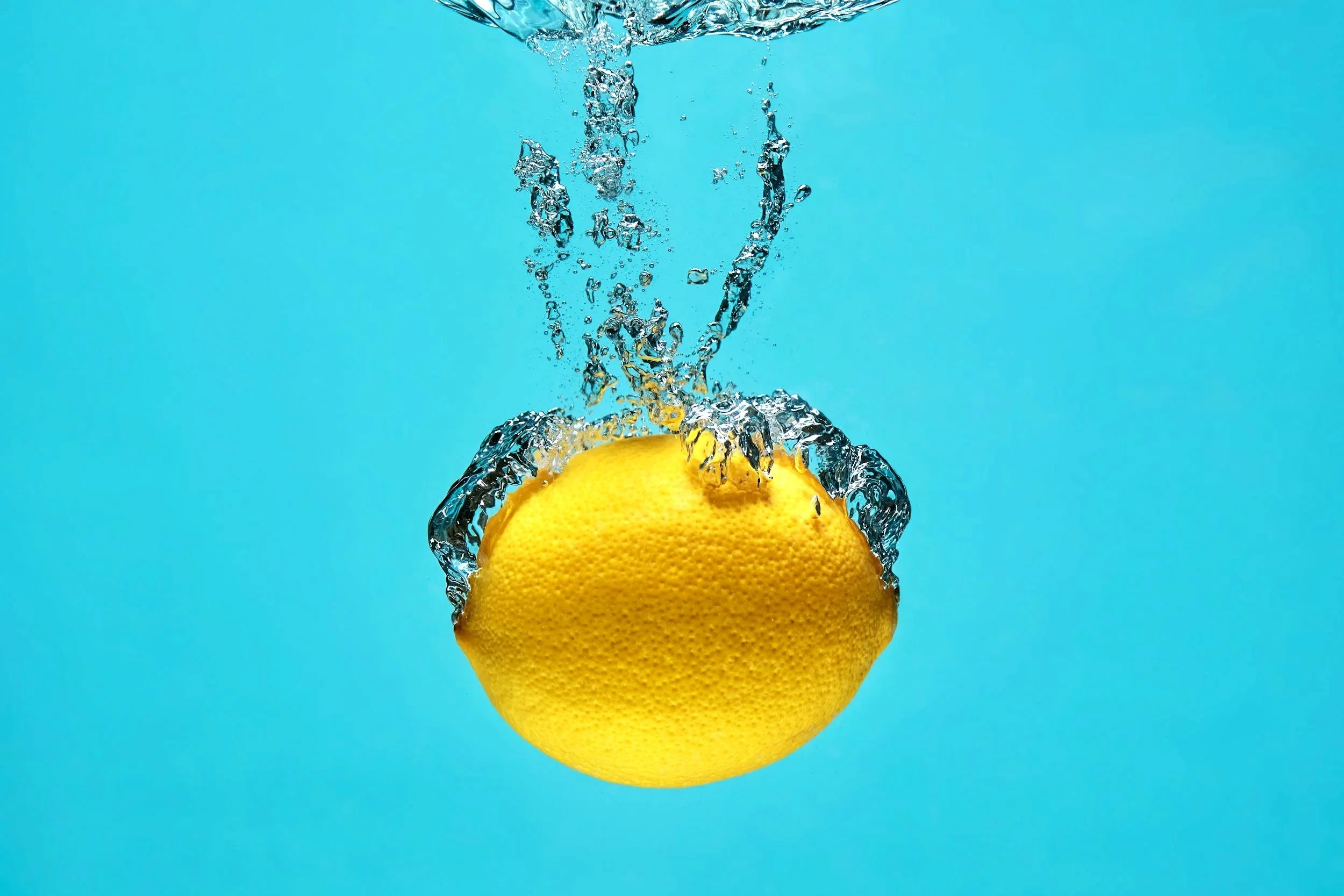 Eine Zitrone taucht in Wasser mit Blasen auf, vor einem hellblauen Hintergrund.