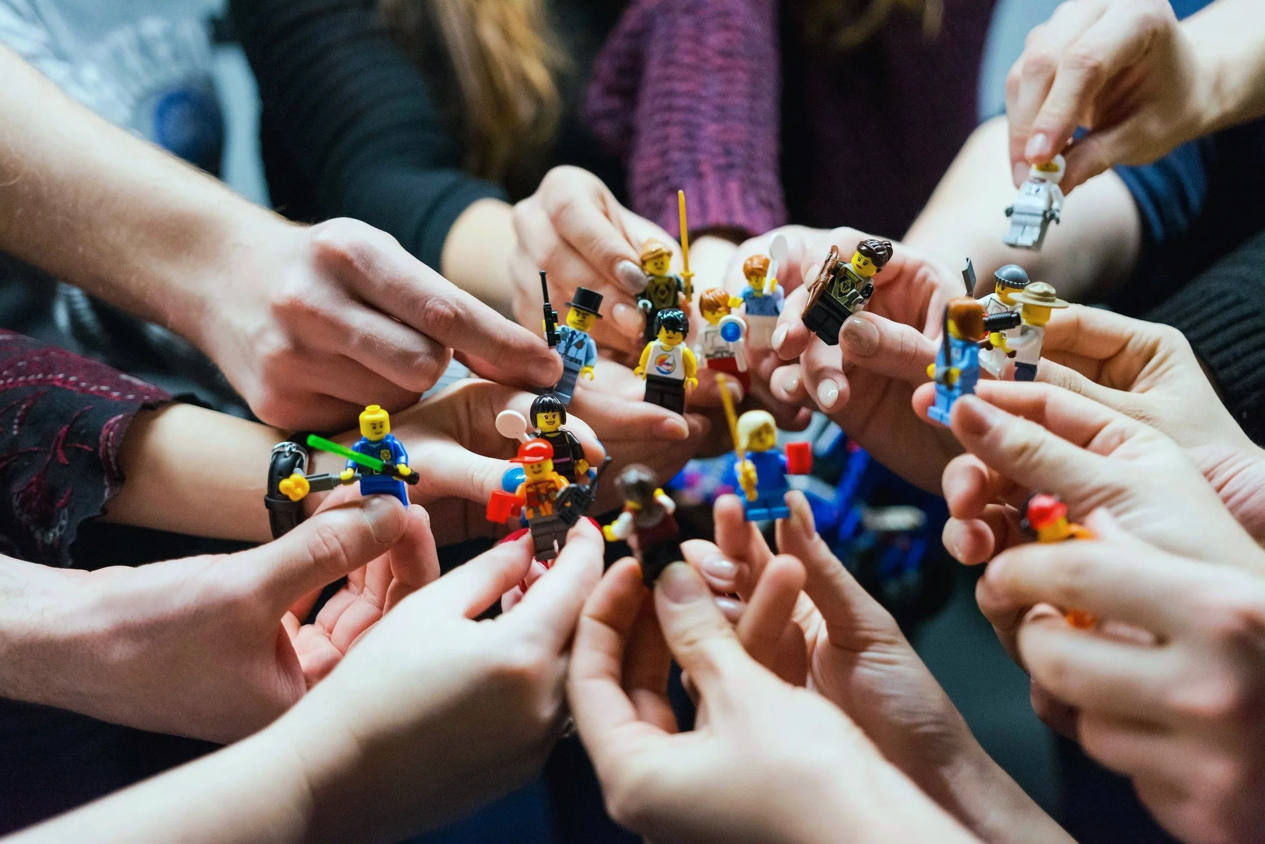 Mehrere Hände, die Lego-Figuren halten und zusammensetzen, im Mittelpunkt eine bunte Sammlung an Lego-Minifiguren als Team.