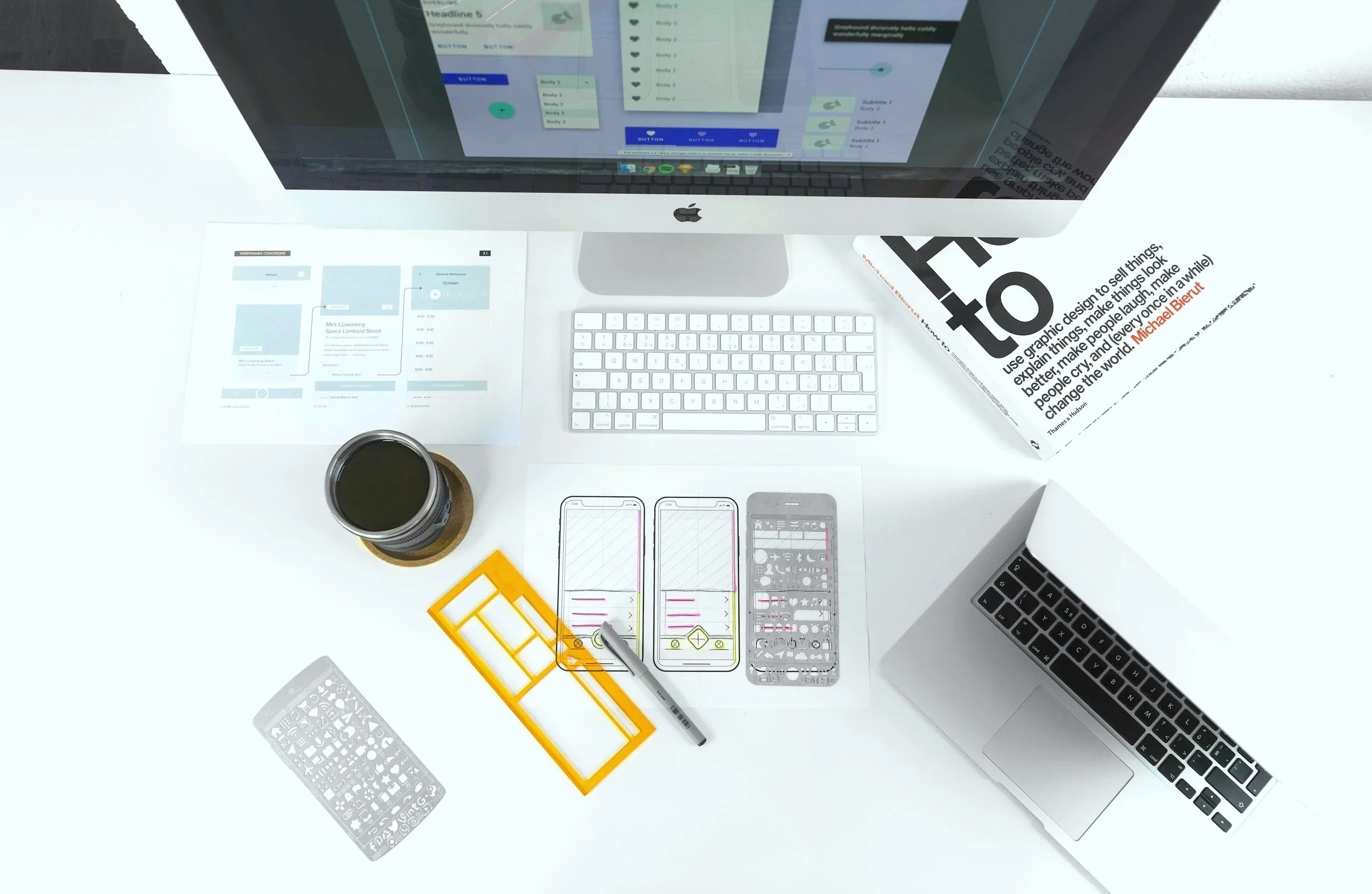 Heller Schreibtisch mit einem iMac, einer weißen Tastatur, einem offenen Laptop, einer gezeichneten Handy-App mit Stift, einem Kaffee, einem Projektplan, einem Booklet, einem orangefarbenen Lineal, einem Papier mit Skizzen und zwei Computermodellen.