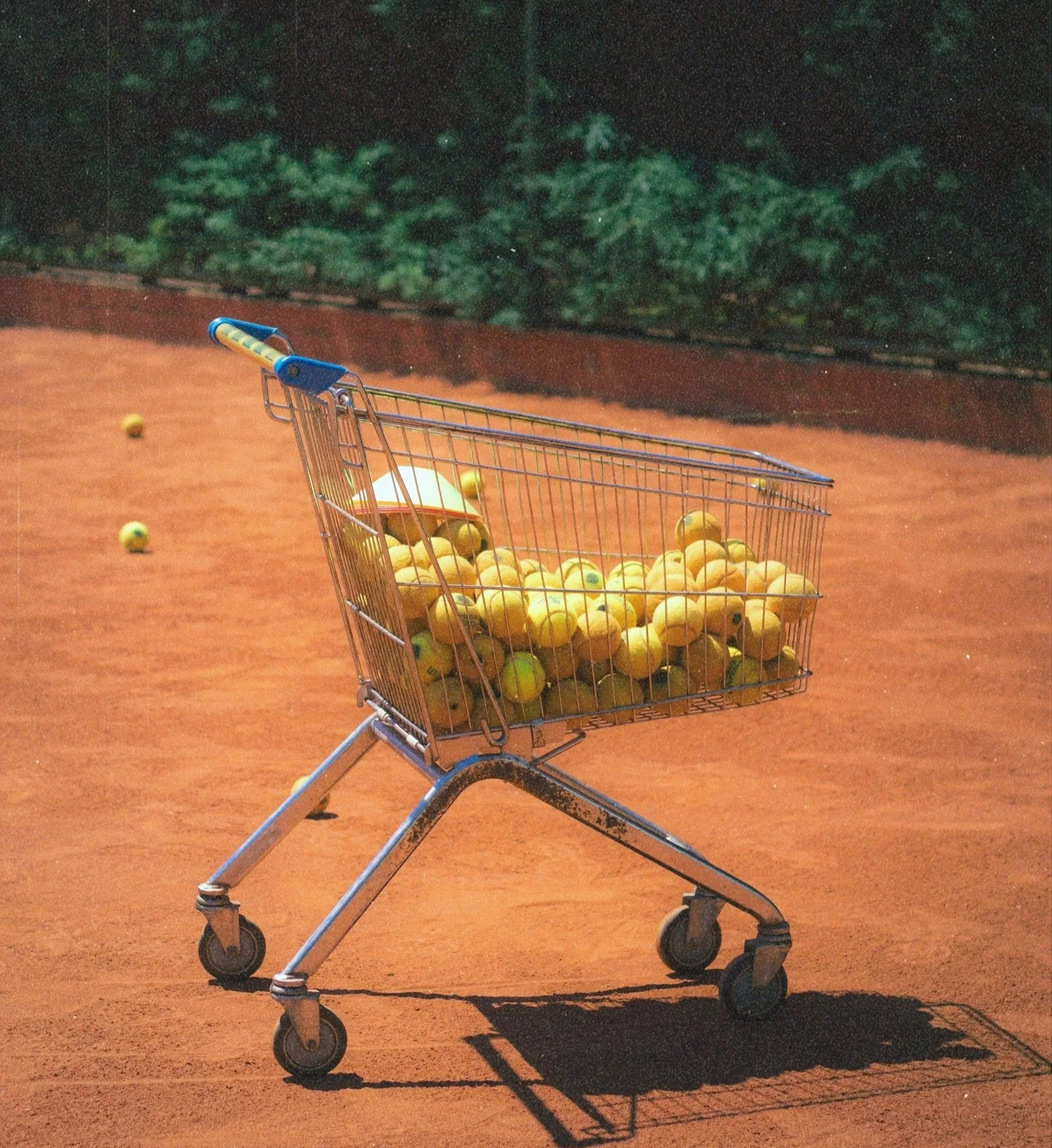 Ein Einkaufswagen auf einem Tennisplatz, gefüllt mit Tennisbällen, einige liegen um den Wagen herum.