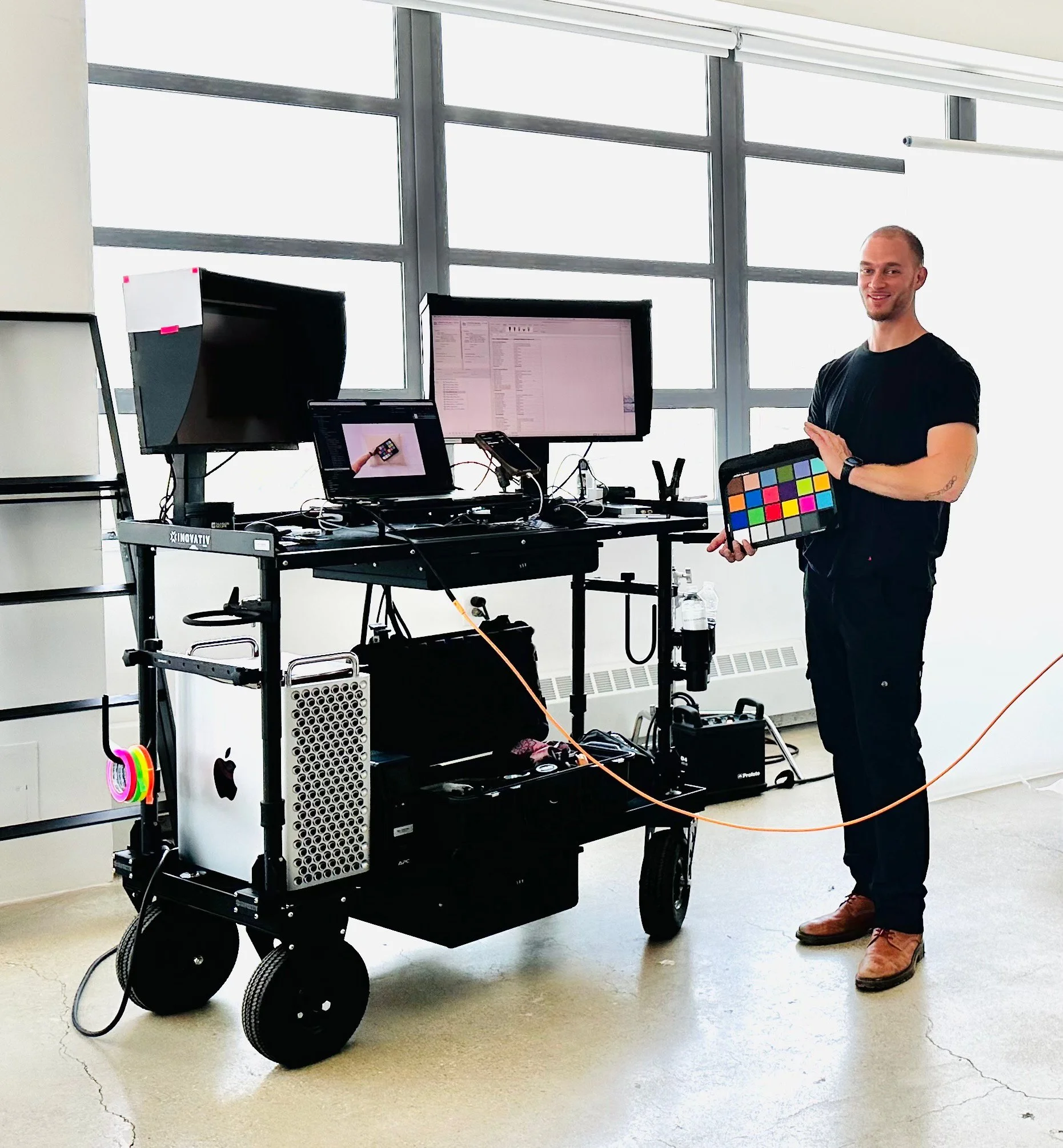 A man standing next to a mobile work station with multiple monitors, holding a color calibration device, in a bright room with large windows.