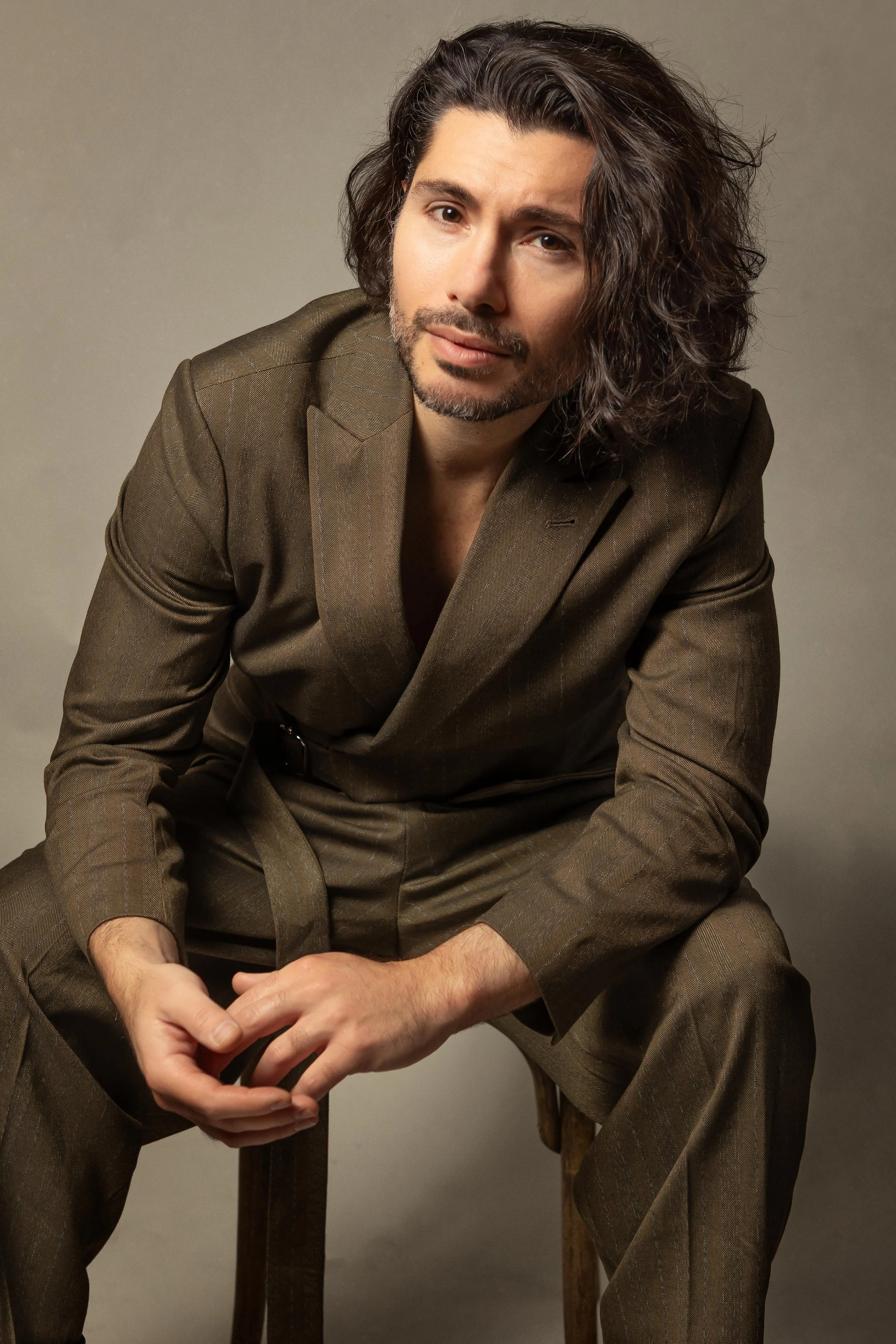 Salvo Maione con lunga chioma riccia, indossa un completo marrone e siede su uno sgabello di legno, guarda verso la fotocamera in modo rilassato.