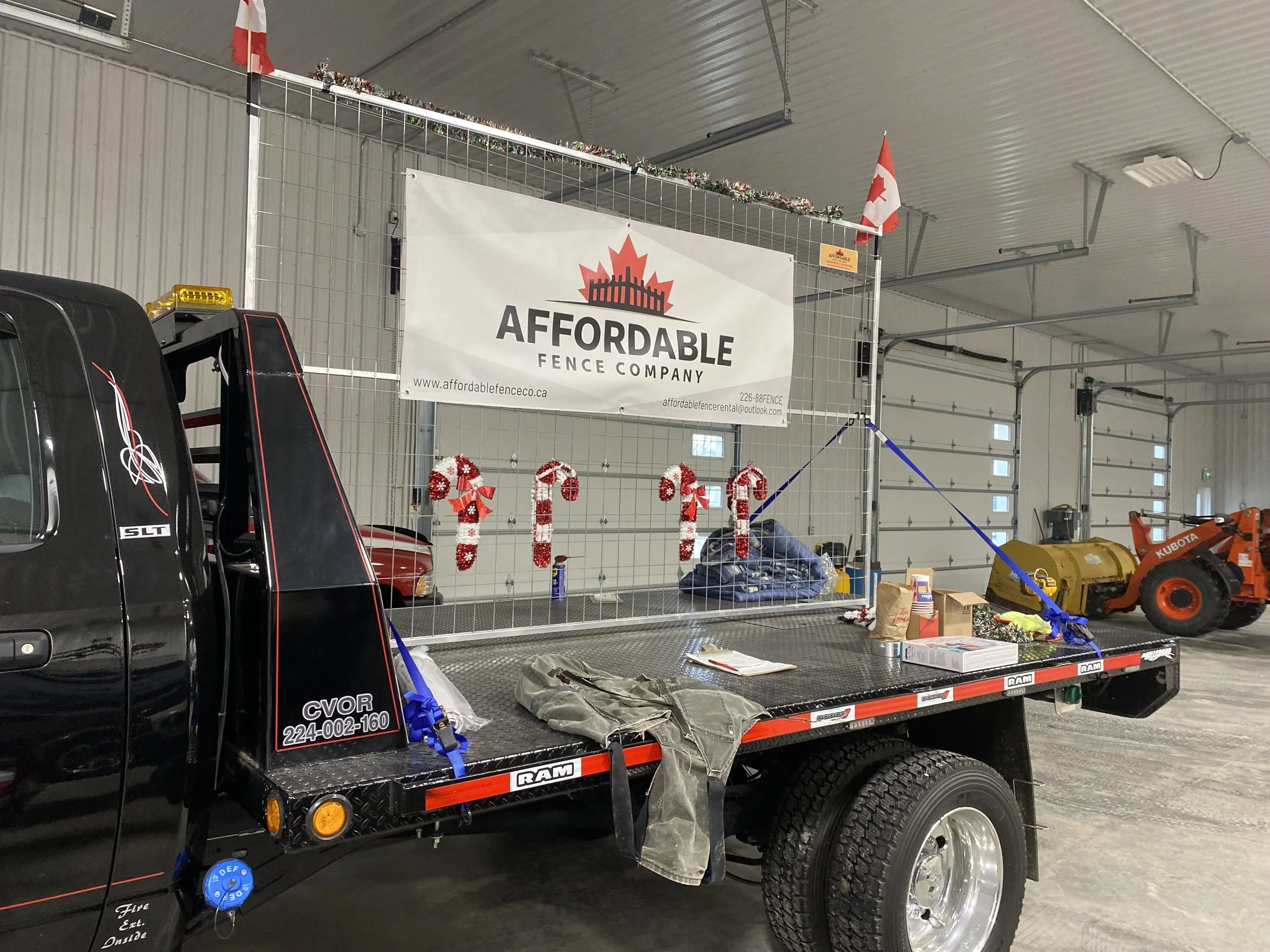 A black flatbed truck with a large sign reading 'Affordable Fence Company' attached to a metal grid, decorated for Christmas with candy cane ornaments and garlands. A Canadian flag is on the top corner of the grid, inside a large industrial garage with a gray metal ceiling and white walls. In the background, there is a yellow skid steer loader, and various tools and boxes on the truck bed.