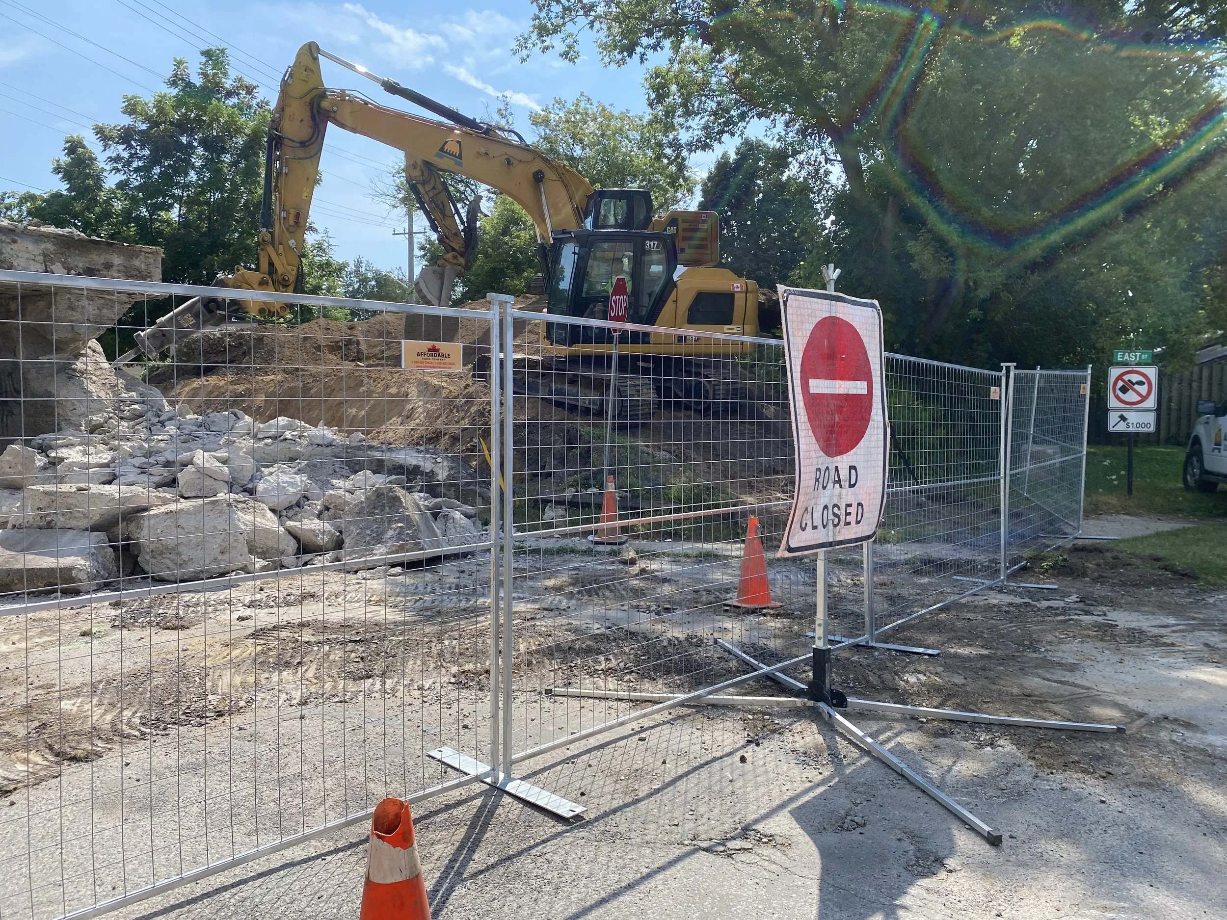 Construction site with an excavator behind a metal fence. A sign indicates the road is closed. Orange cones are on the ground. There are trees and signs in the background.