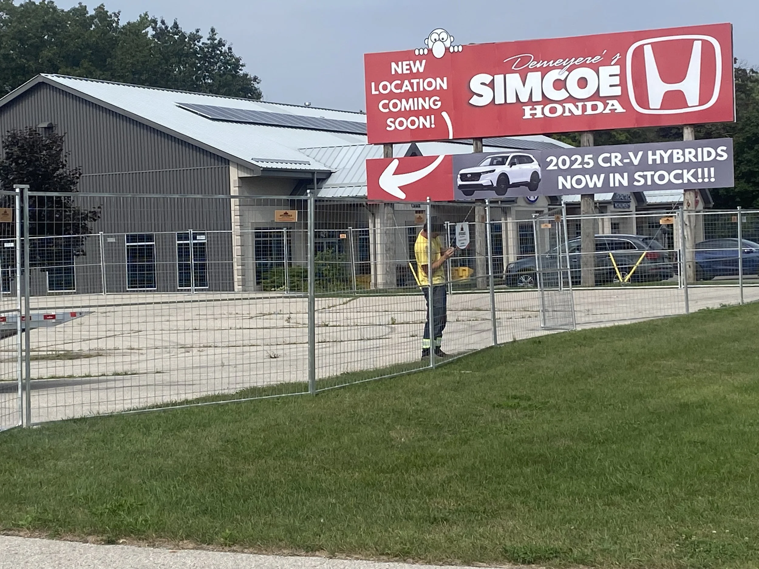 Large red and white billboard advertising a new location of Simcoe Honda, featuring a cartoon face at the top. It mentions the arrival of the 2025 CR-V Hybrids in stock, with an arrow pointing to the dealership behind the fence, which has a building with solar panels on the roof. A person in a yellow shirt appears to be working or inspecting near the fence.