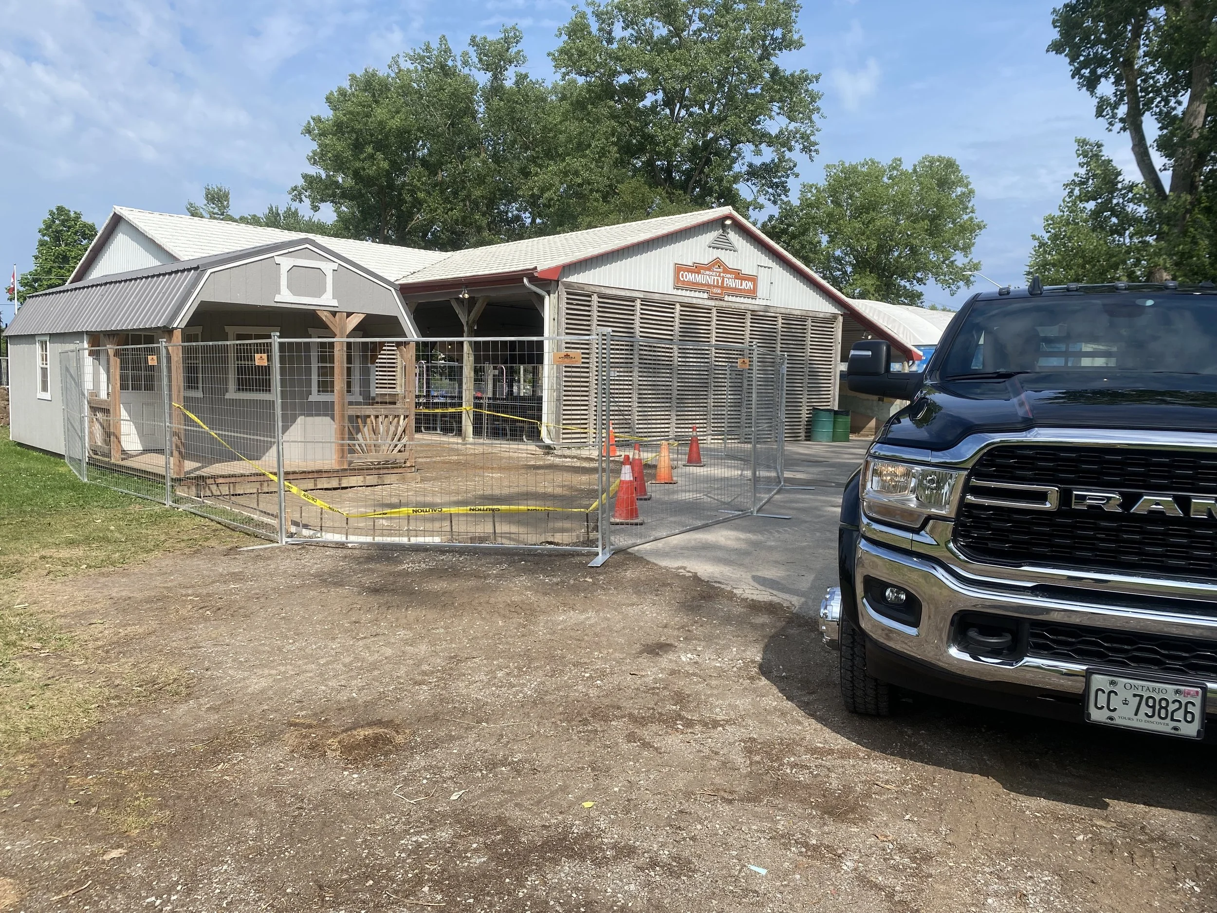 Community pavilion under construction, surrounded by a metal fence with caution tape, orange traffic cones, and a black Ram truck parked nearby.